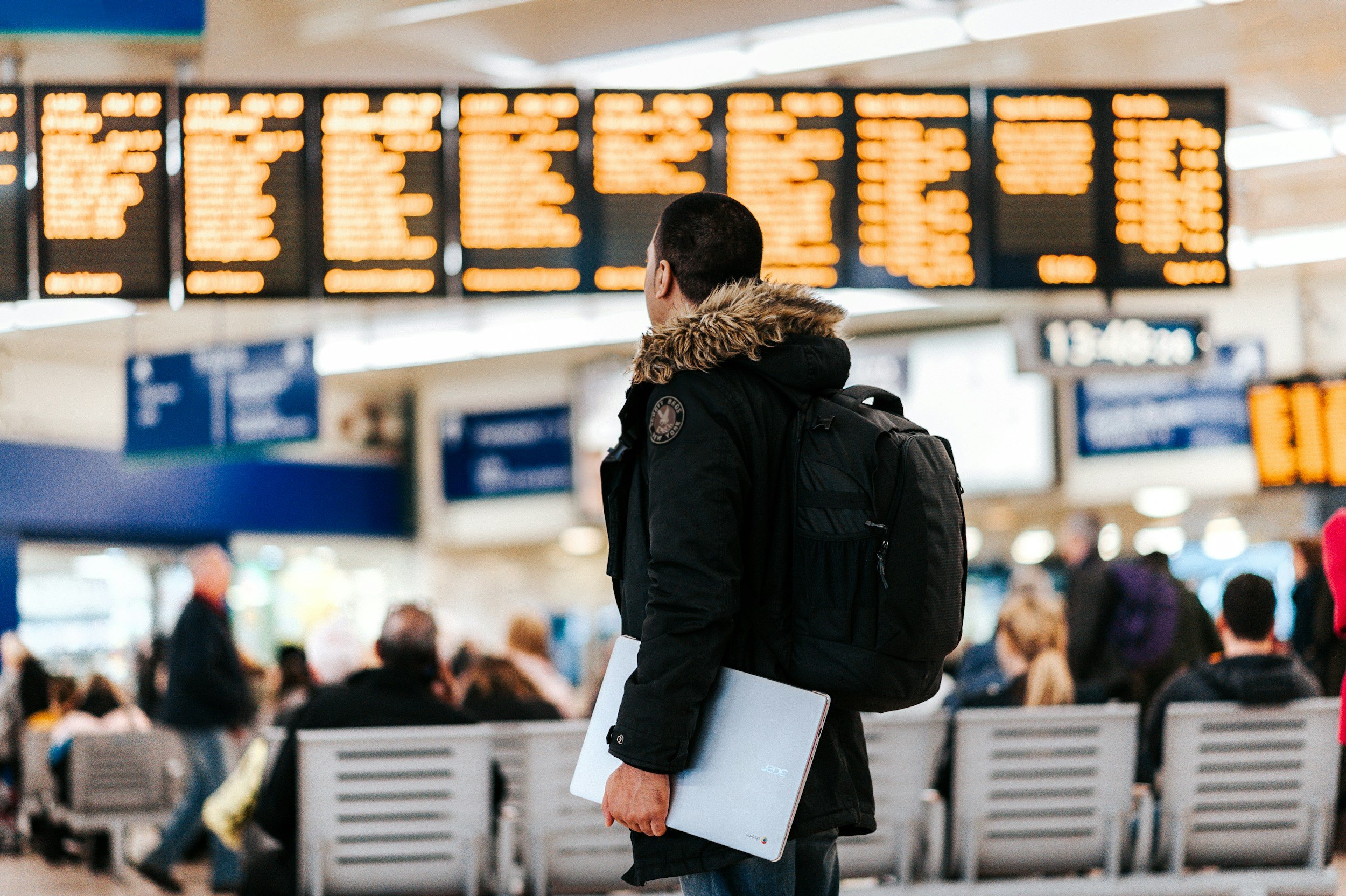 A person with short dark hair, wearing a black winter coat with a fur-lined hood and a backpack, holding a silver laptop, standing in a busy airport terminal in front of the flight information display board.