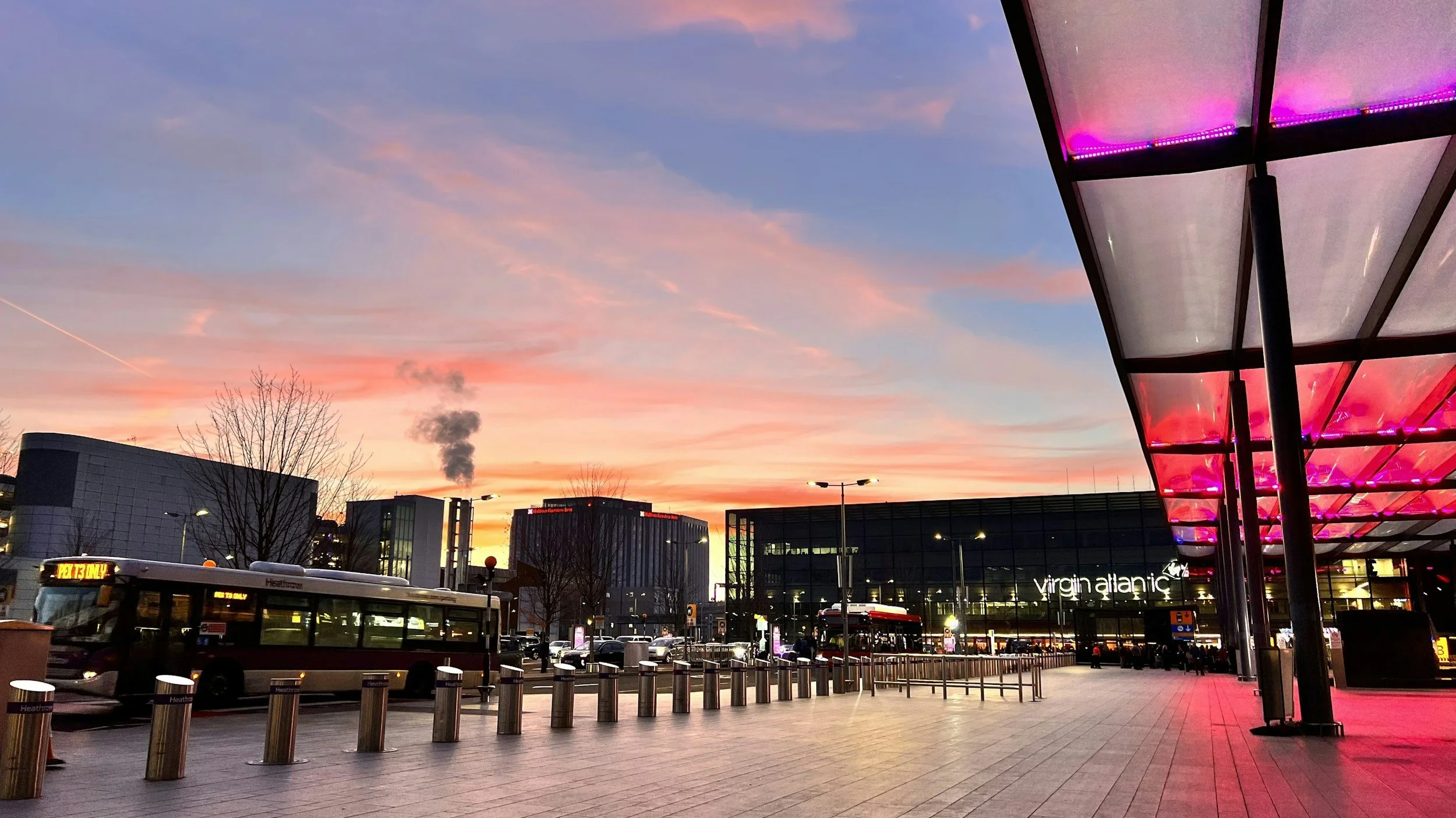 A cityscape at sunset with colorful sky, modern buildings, and Virgin Atlantic sign on a building, with buses and pedestrians in the foreground.