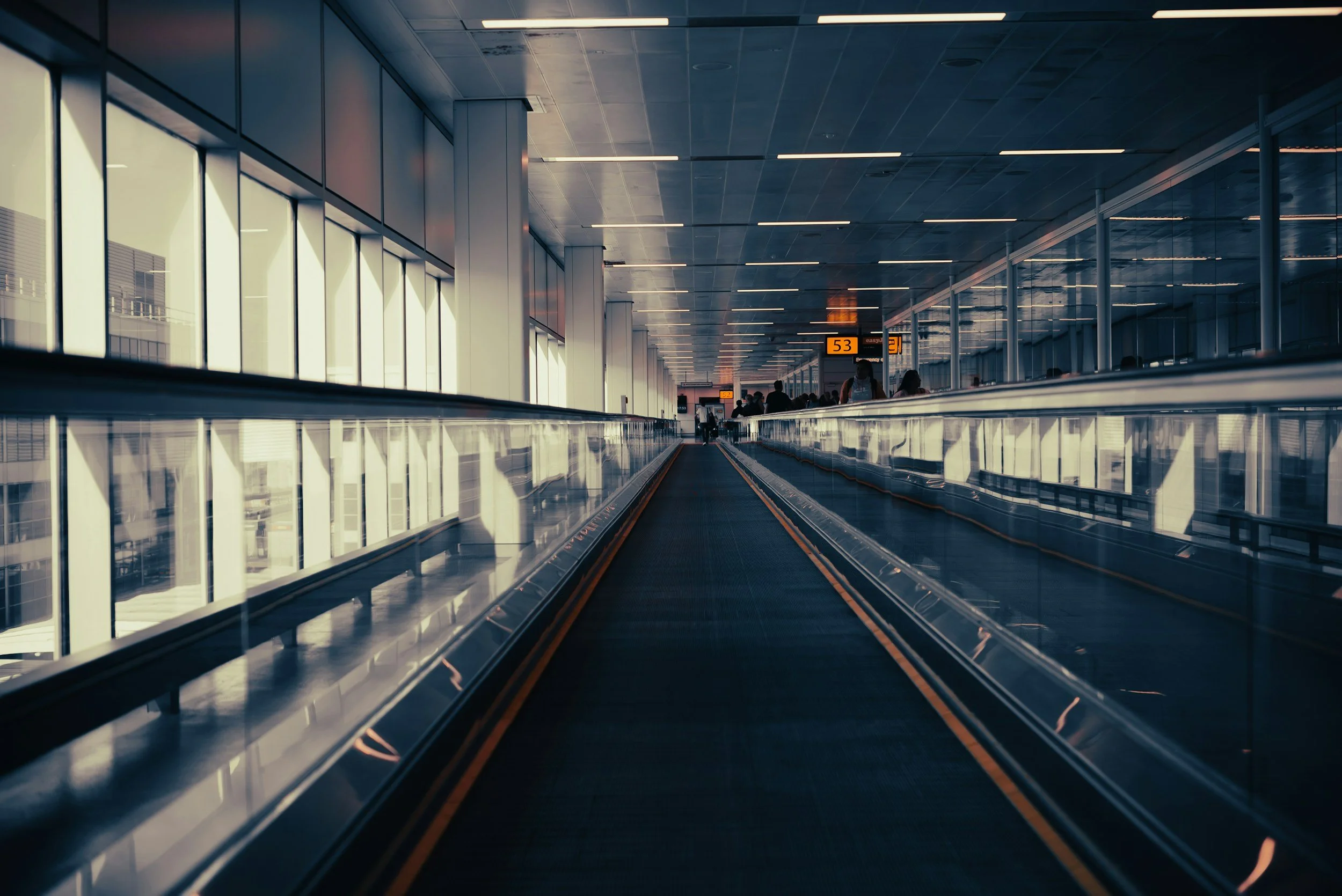 Indoor airport terminal with moving walkways, large windows on the left, and passengers walking in the background, with digital flight information signs hanging from the ceiling.