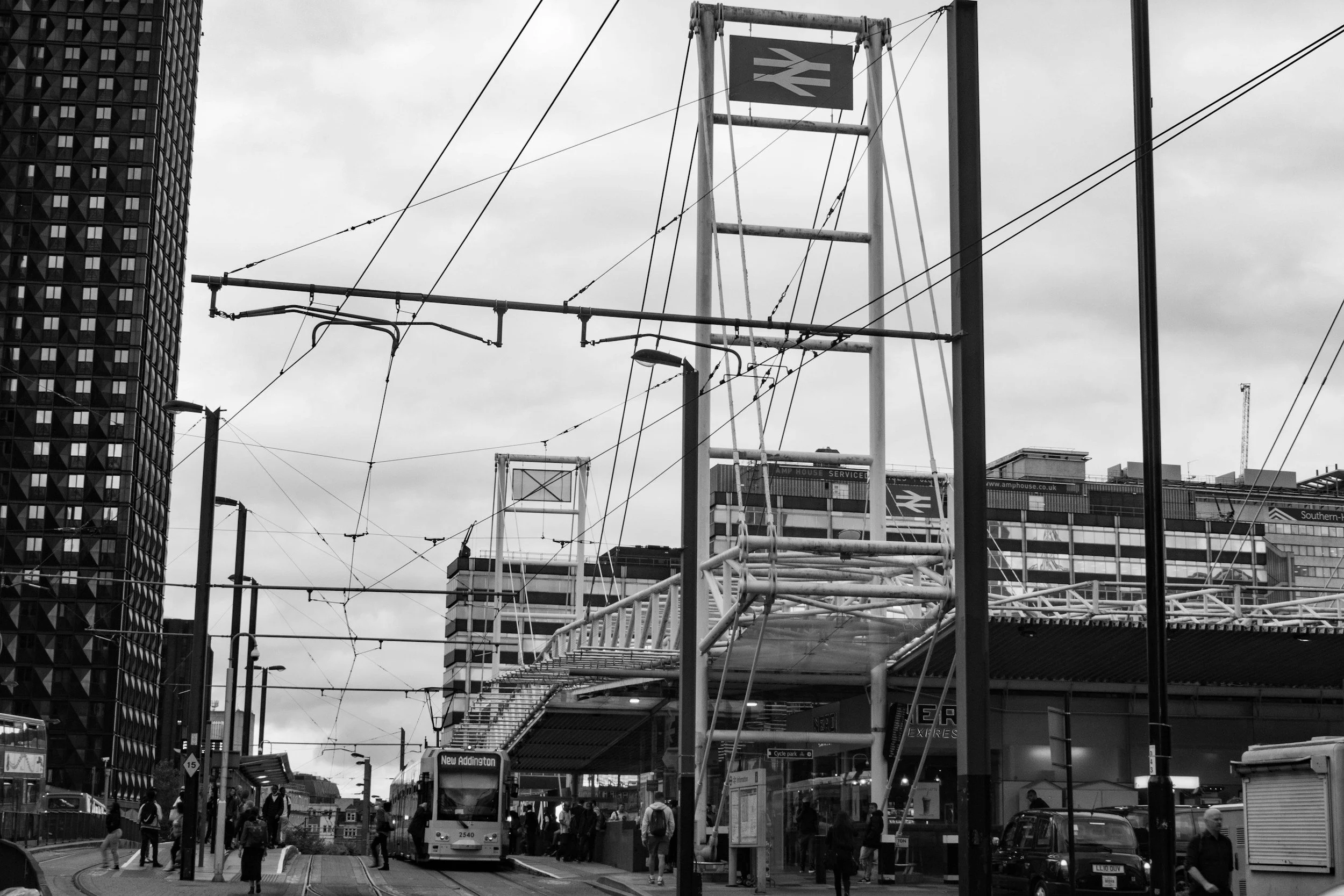 City train station with a train and people walking on the platform, overhead electrical wires, and skyscrapers in the background.