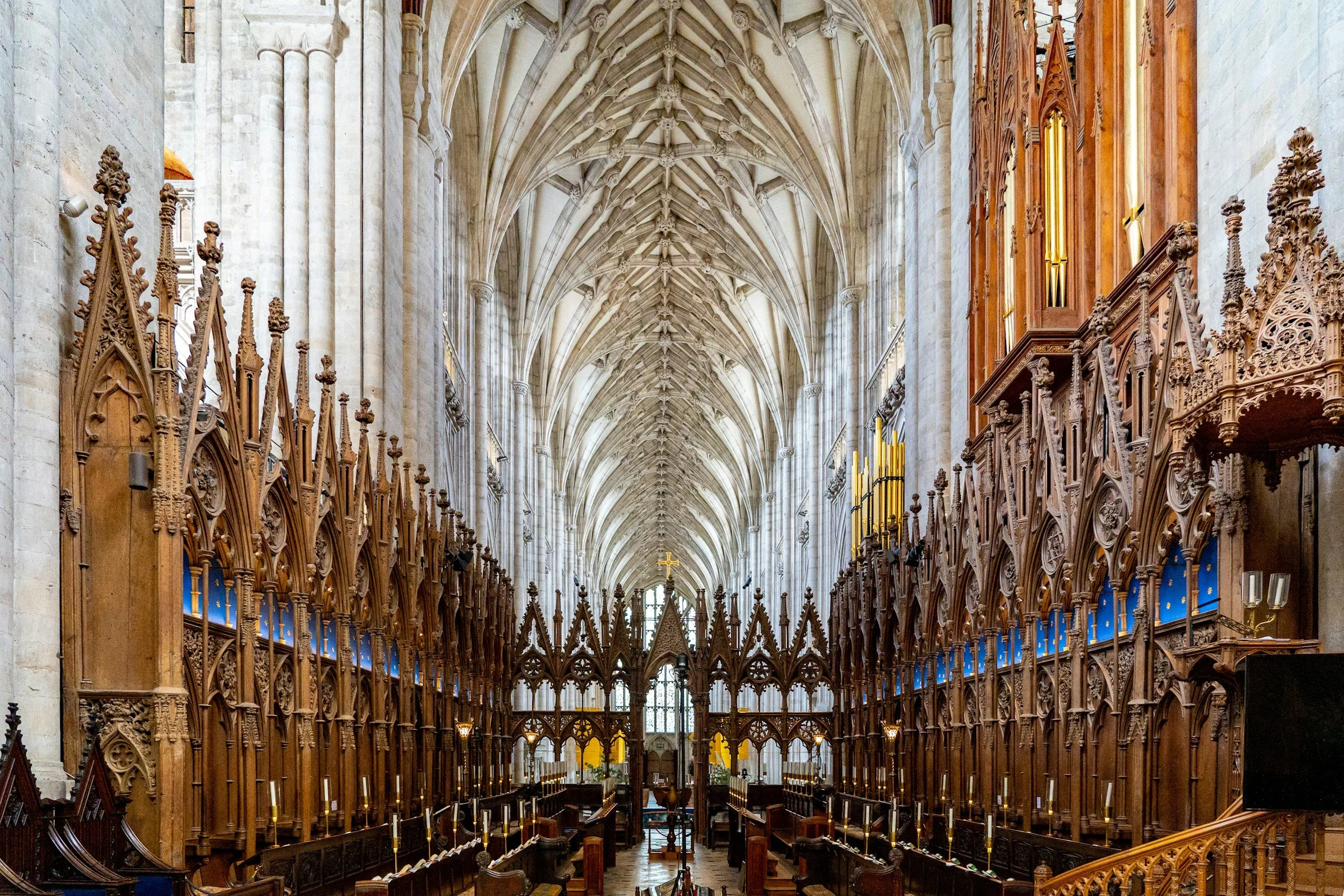 Interior view of a Gothic cathedral with high vaulted ceilings, intricate wooden choir stalls, and sunlight streaming through stained glass windows.