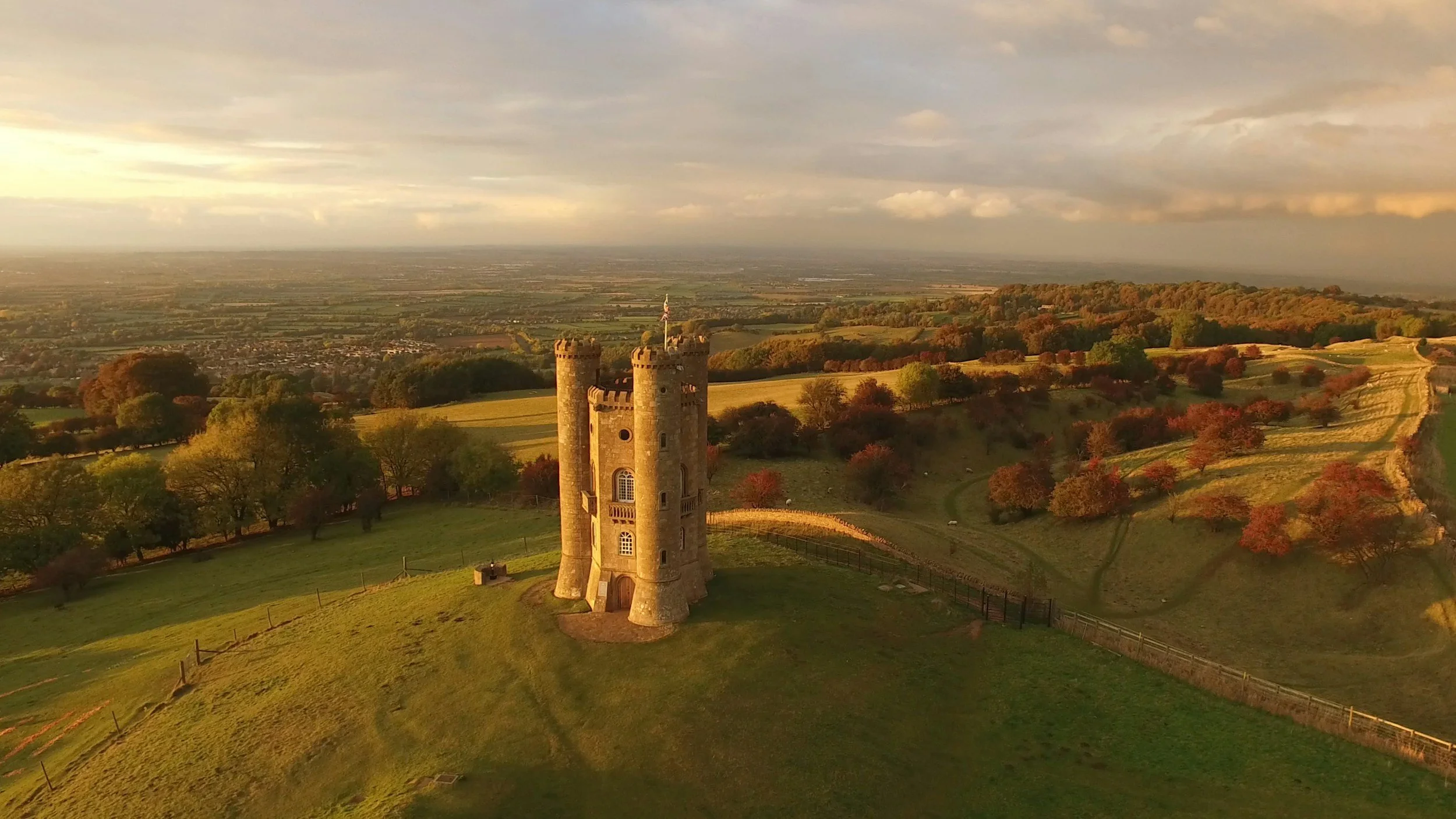 A scenic view of a castle tower surrounded by rolling hills with colorful autumn trees and a vast landscape in the background, under a partly cloudy sky.