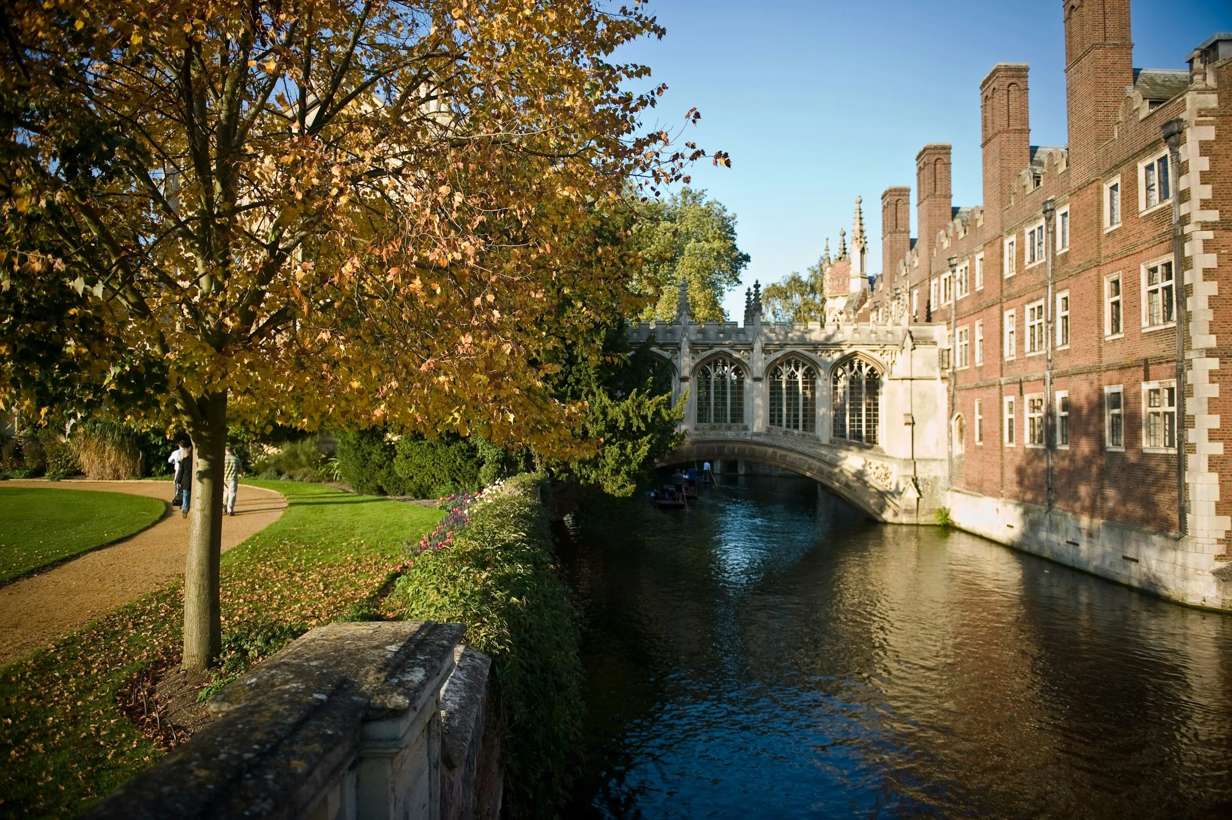 A scenic view of a historic brick building with multiple chimneys, a stone bridge over a canal, a large tree with autumn-colored leaves, and a grassy park with people walking along a curved path.