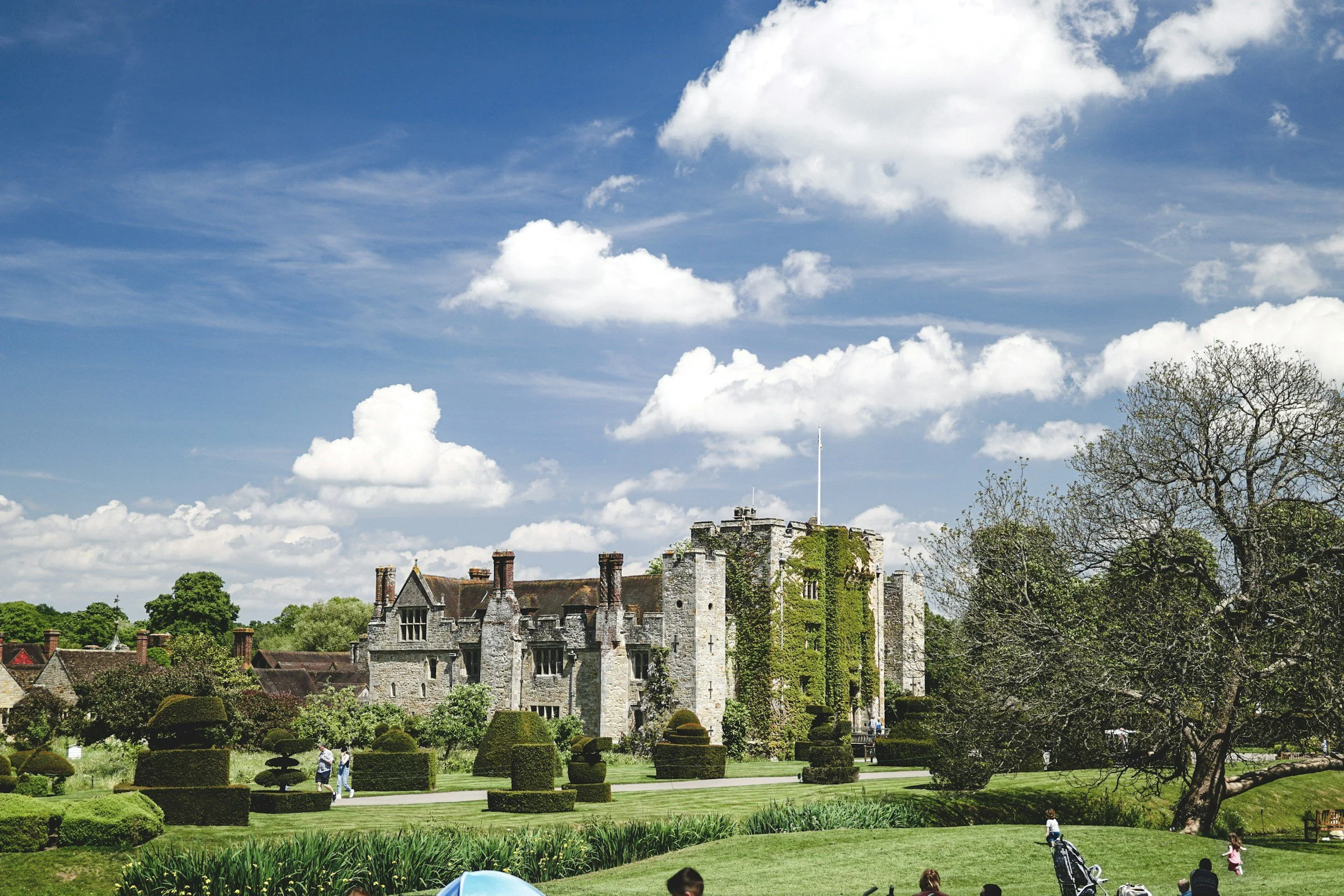 A historic stone castle with turrets and crenellations, surrounded by a well-manicured garden and lush trees, under a partly cloudy blue sky.