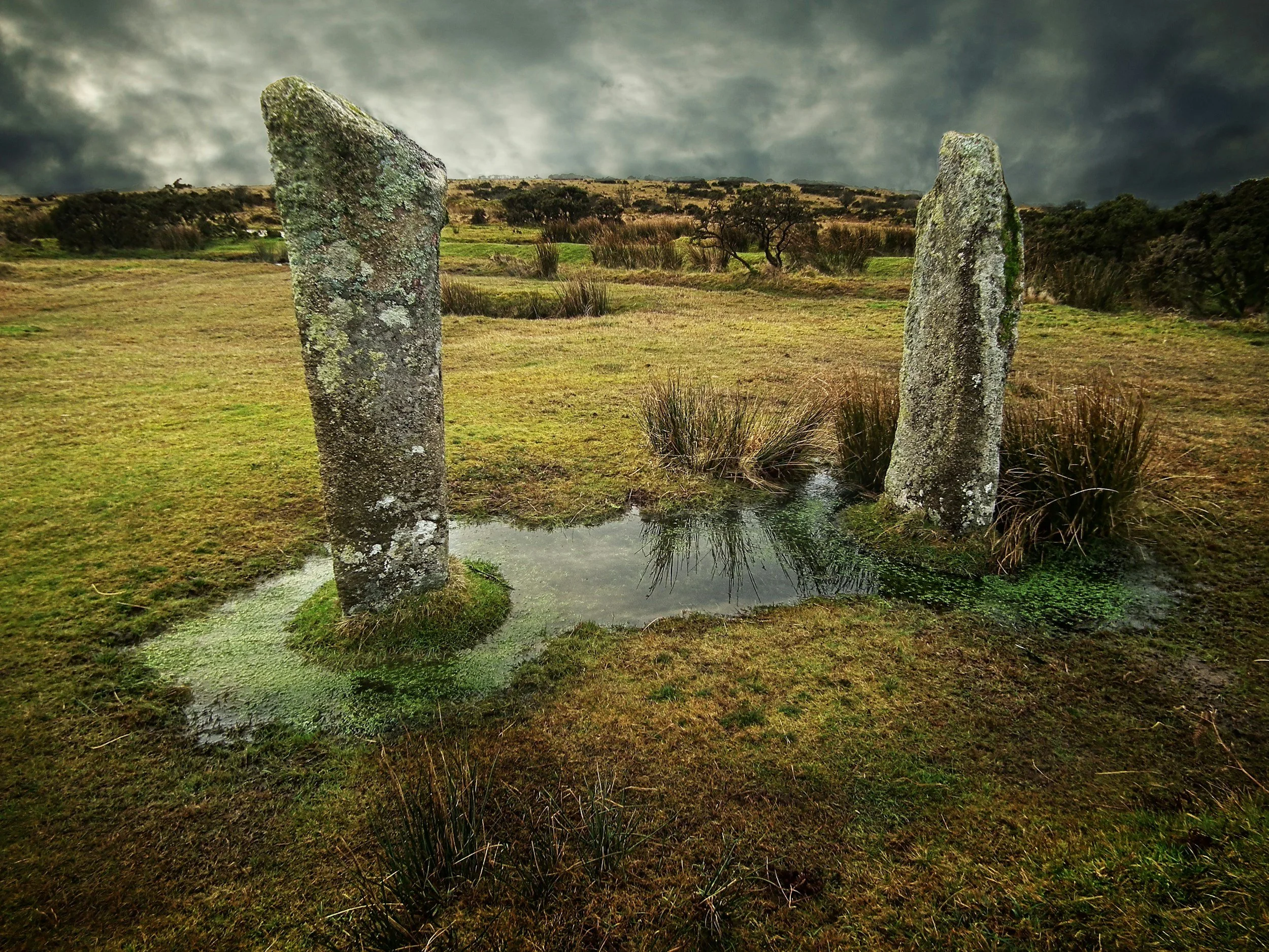Two ancient standing stones in a grassy landscape with rolling hills and a cloudy sky in the background.