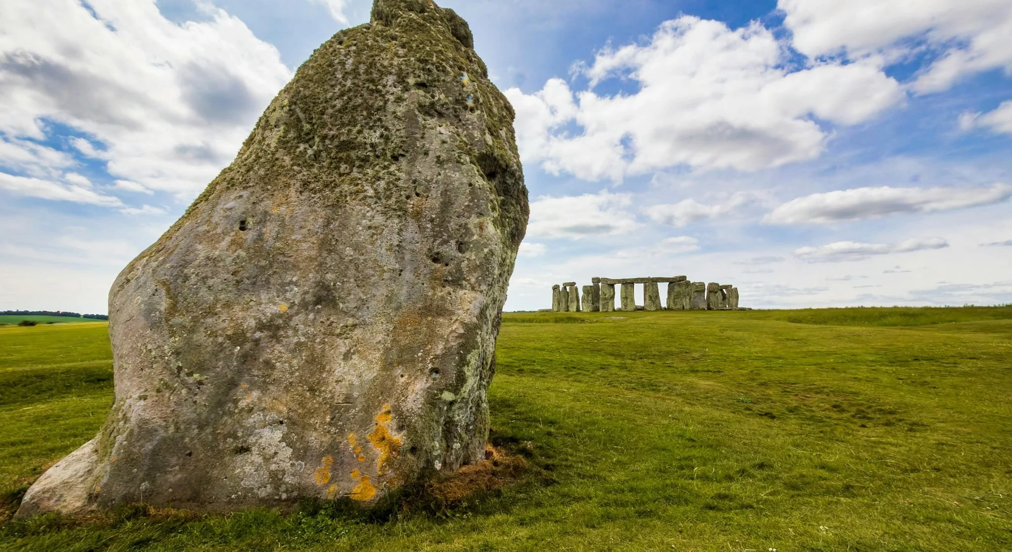 View of Stonehenge with a large weathered stone in the foreground and the iconic stone circle in the background, under a partly cloudy sky.