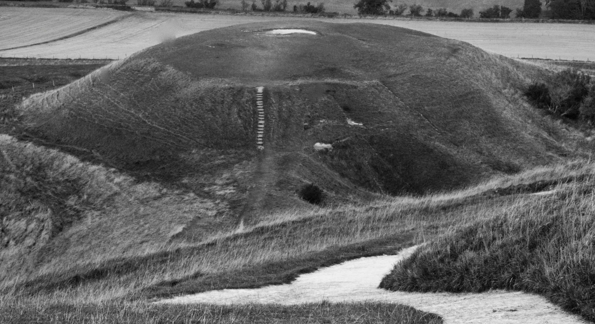 A large, rounded grass-covered hill with a small white circular area at the top, surrounded by open fields and patches of grass.