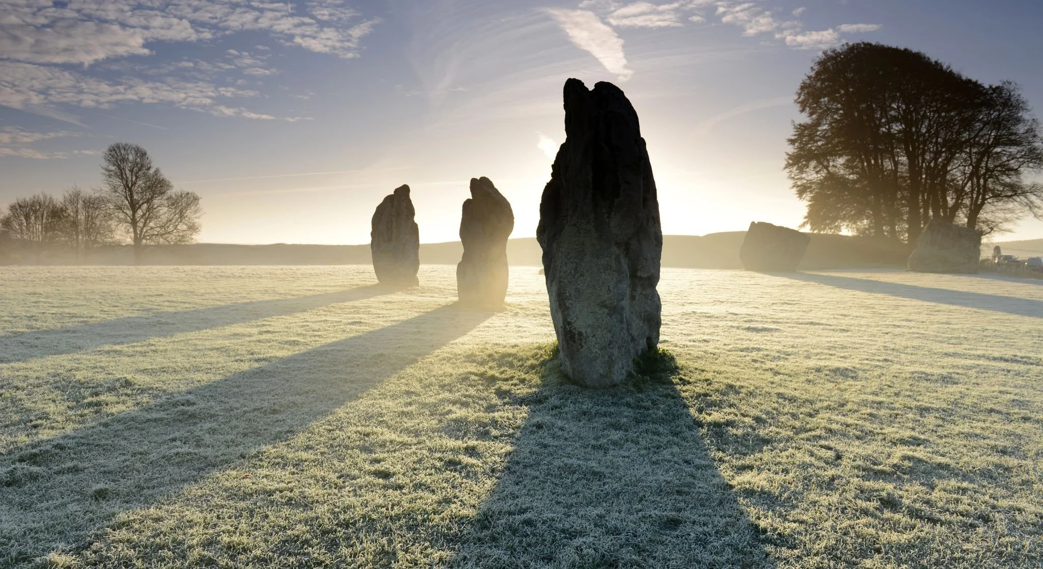 Sunrise over a grassy field with standing stones, casting long shadows. Trees are visible in the background, and a slight mist or frost covers the ground.