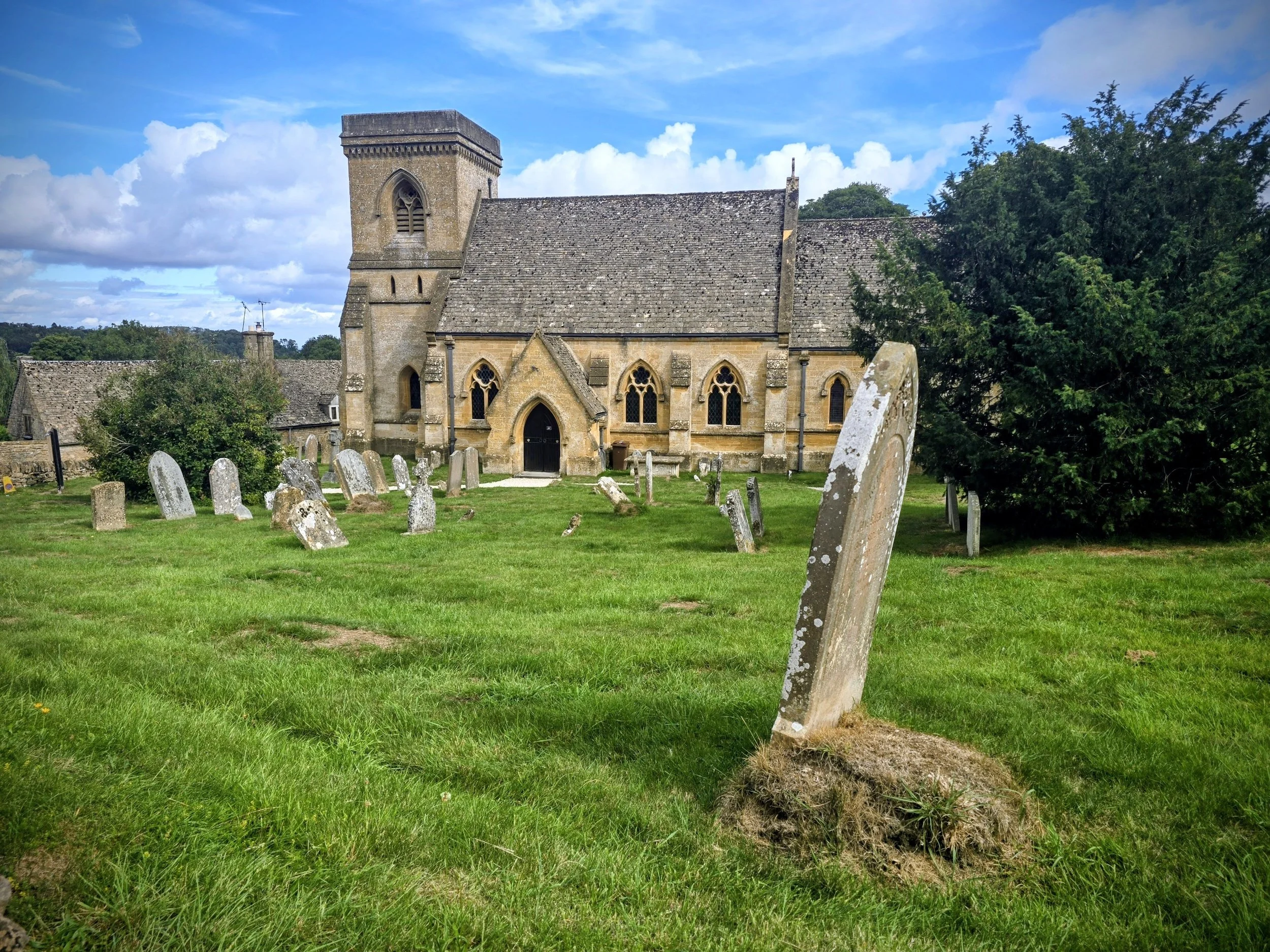 A historic stone church with a graveyard in front, surrounded by green grass, trees, and a blue sky with clouds.