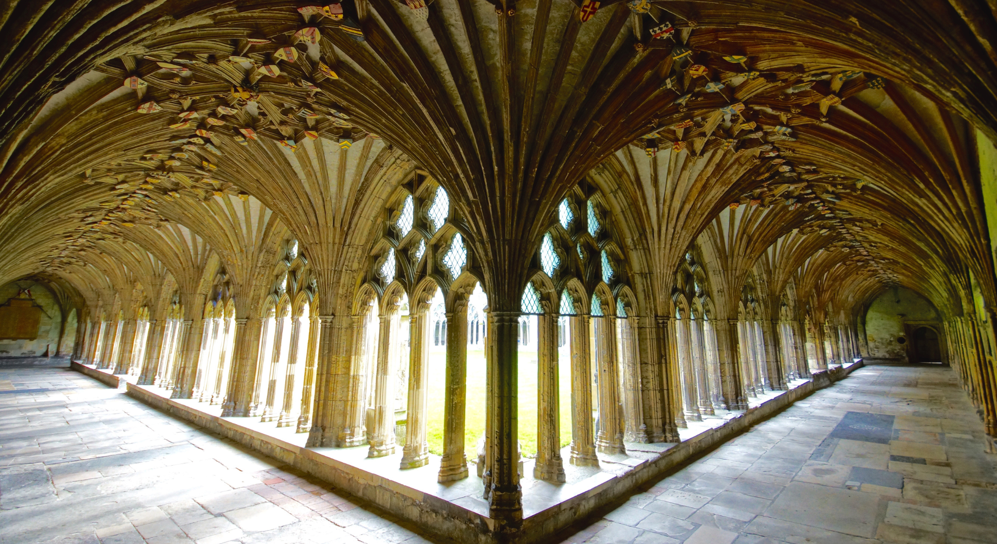 View of an ancient stone cloister with pointed archways and high vaulted wooden ceiling, illuminated by natural light through windows with diamond-shaped panes.