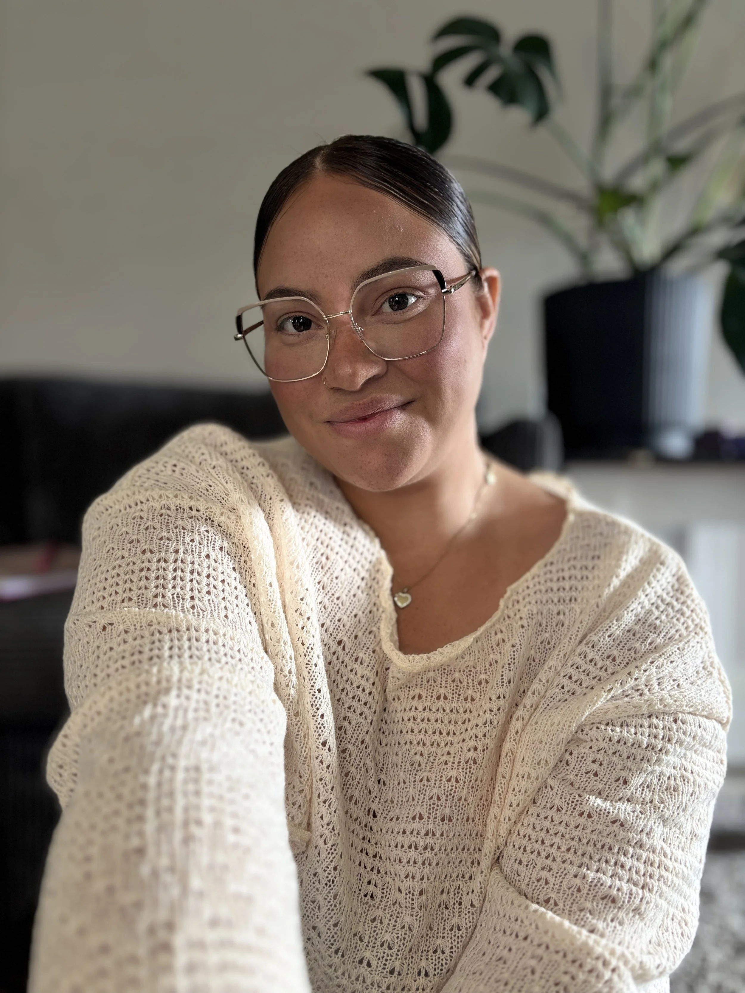 A woman with glasses and a smile taking a selfie in a room with a black couch, a plant, and window blinds in the background.