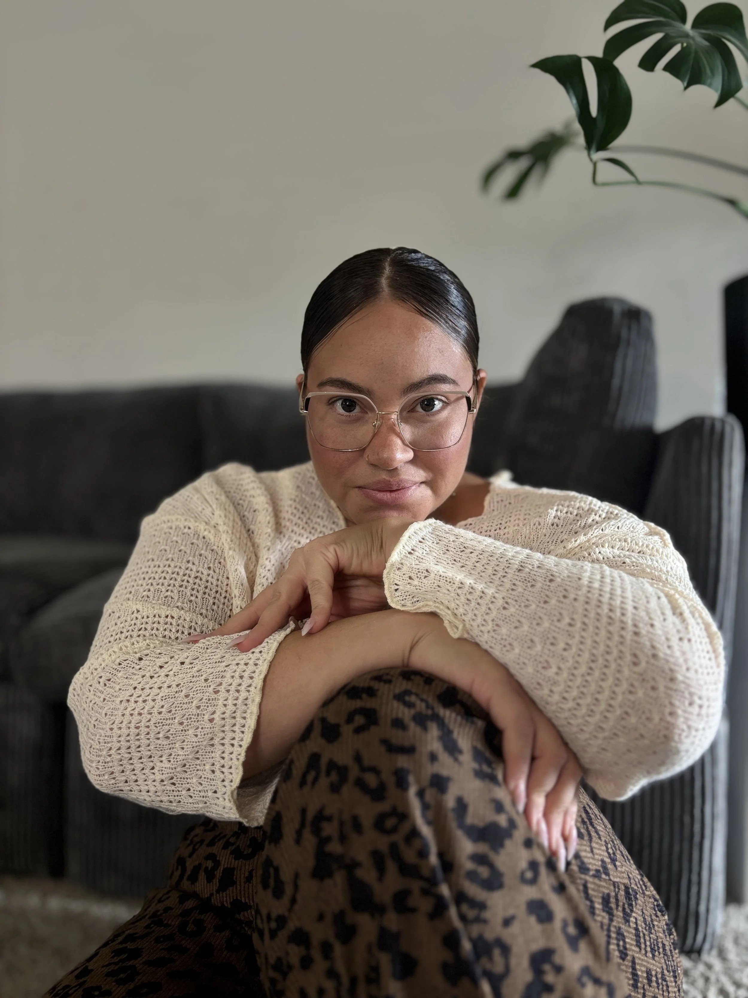 Woman with glasses sitting on a couch, resting her arms on her knee, looking at the camera with a neutral expression.
