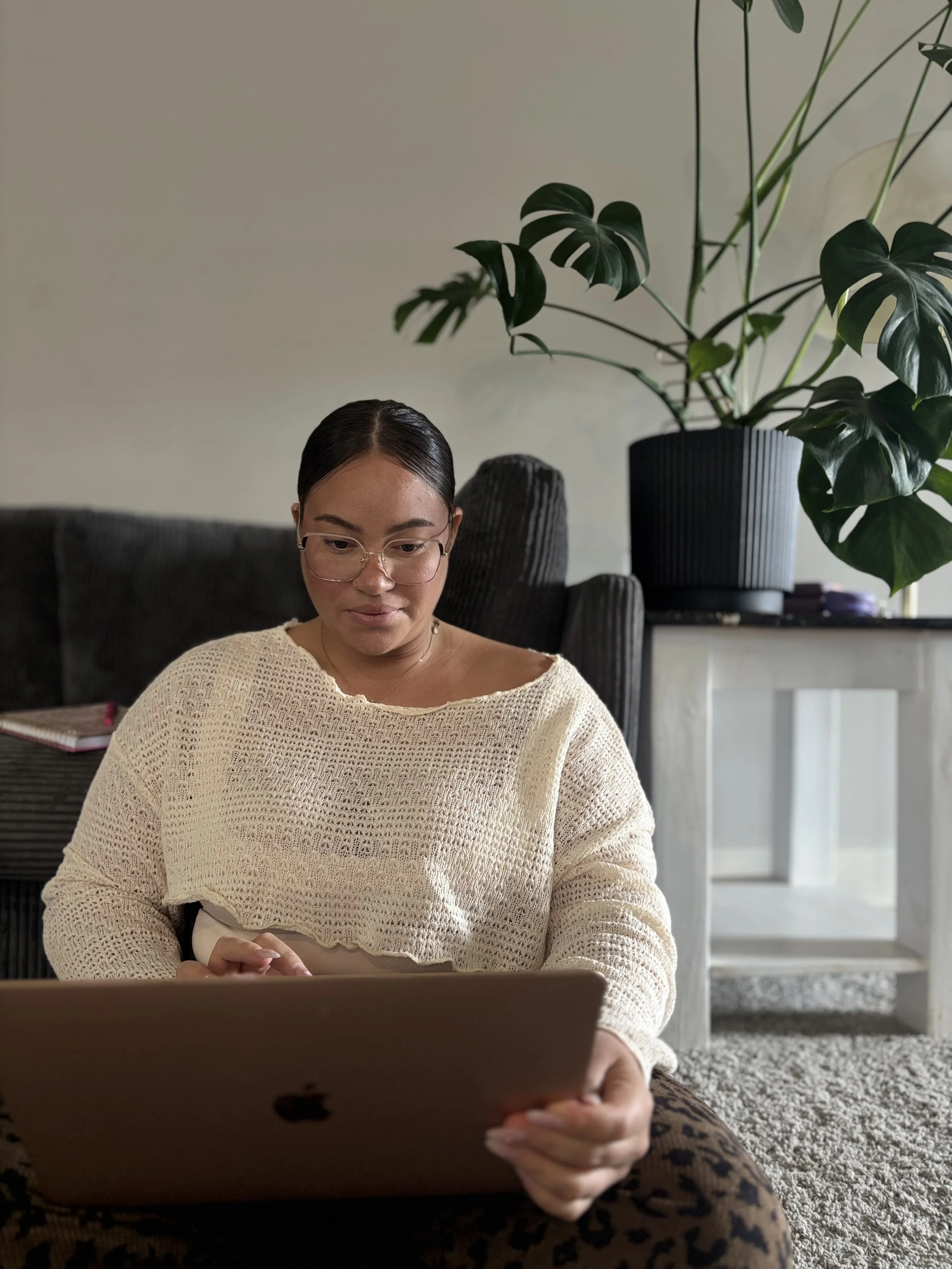 Woman sitting on the floor using a laptop in a living room with a large plant behind her.