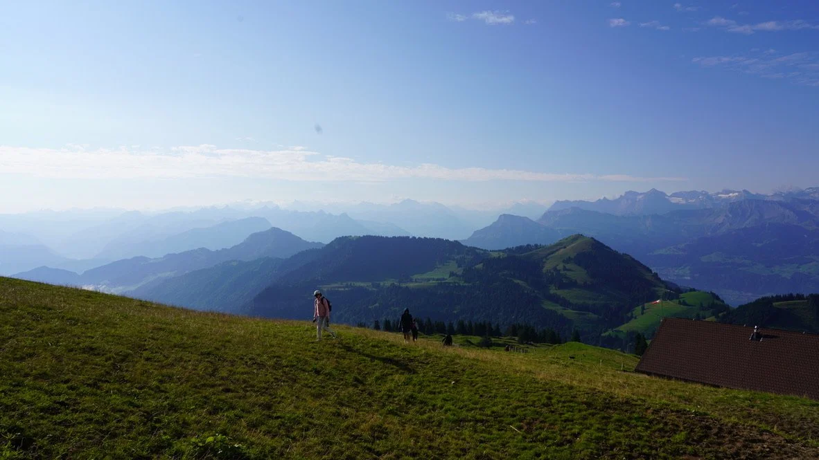 Hikers walking on a grassy hillside with mountains in the background and a partly cloudy sky.
