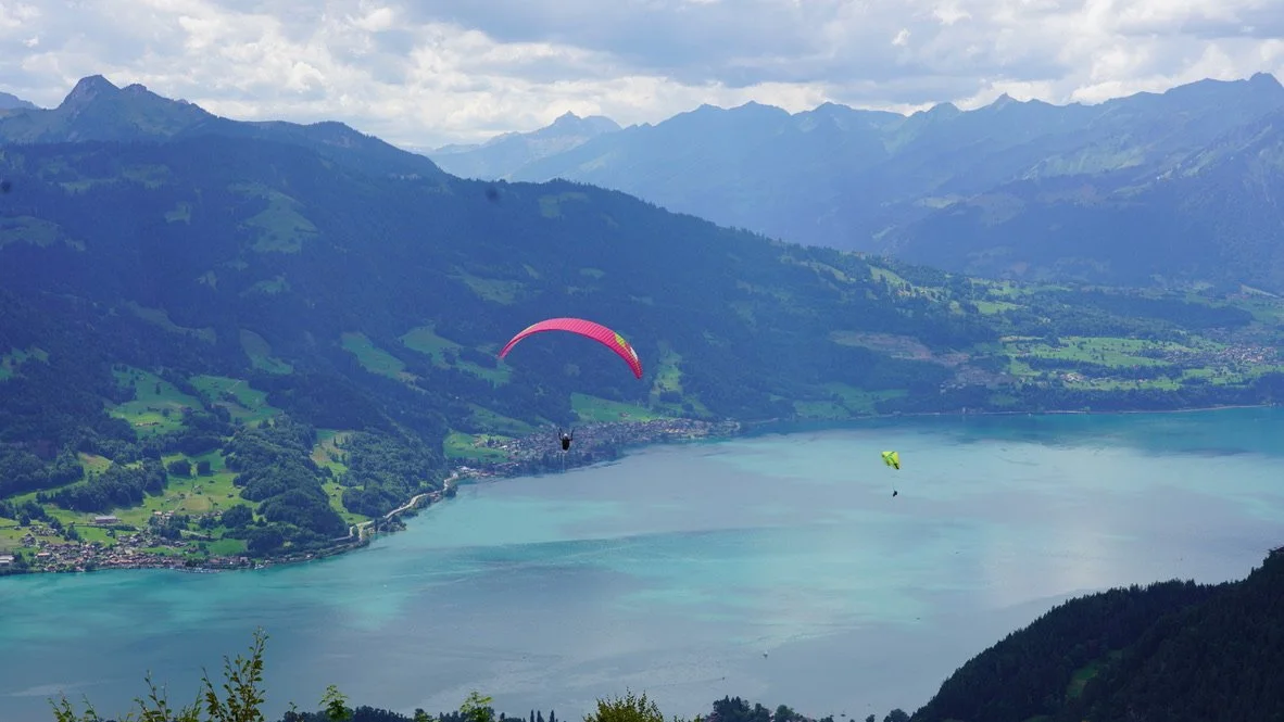Two paragliders flying over a large, calm lake surrounded by green mountains.