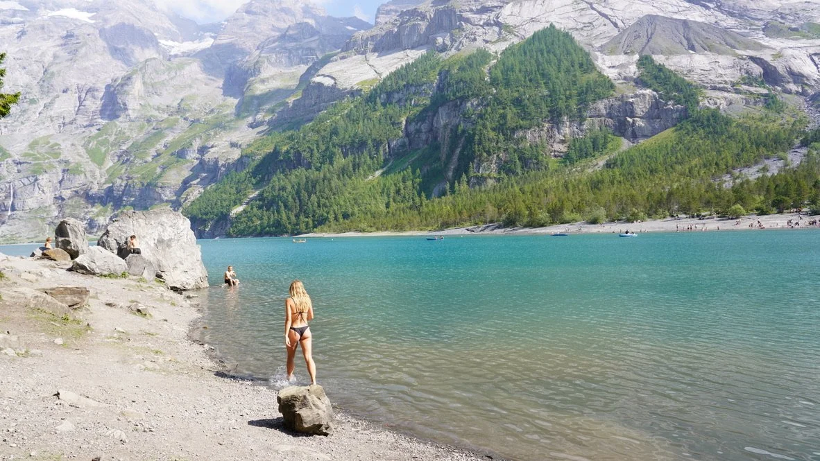 A woman in a swimsuit standing on a rock at the edge of a lake with mountains and green forest in the background.