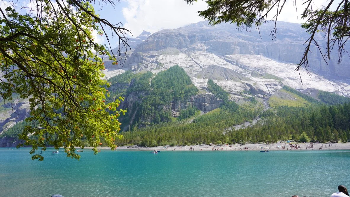 Scenic view of a mountain lake with turquoise water, surrounded by green trees and rocky mountain slopes in the background, with people on the shoreline.