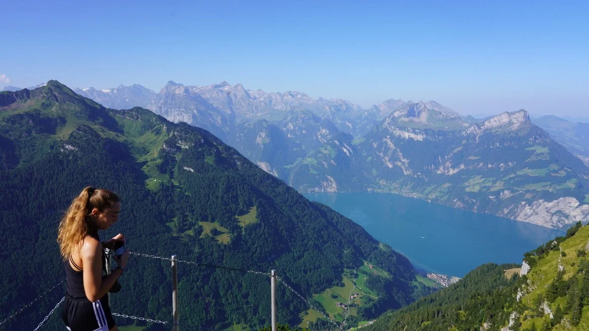 A woman standing on a mountain trail overlooks a lake and mountain range in a scenic landscape.