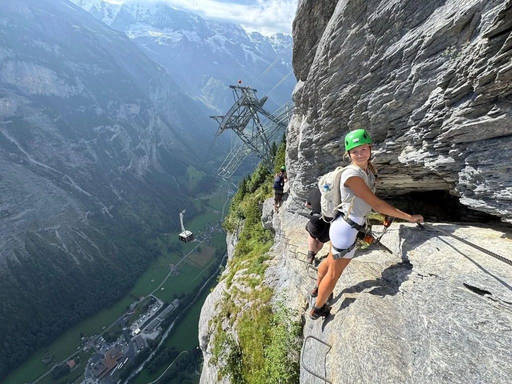 People rock climbing on a steep mountain face with a view of a valley and mountains in the background.