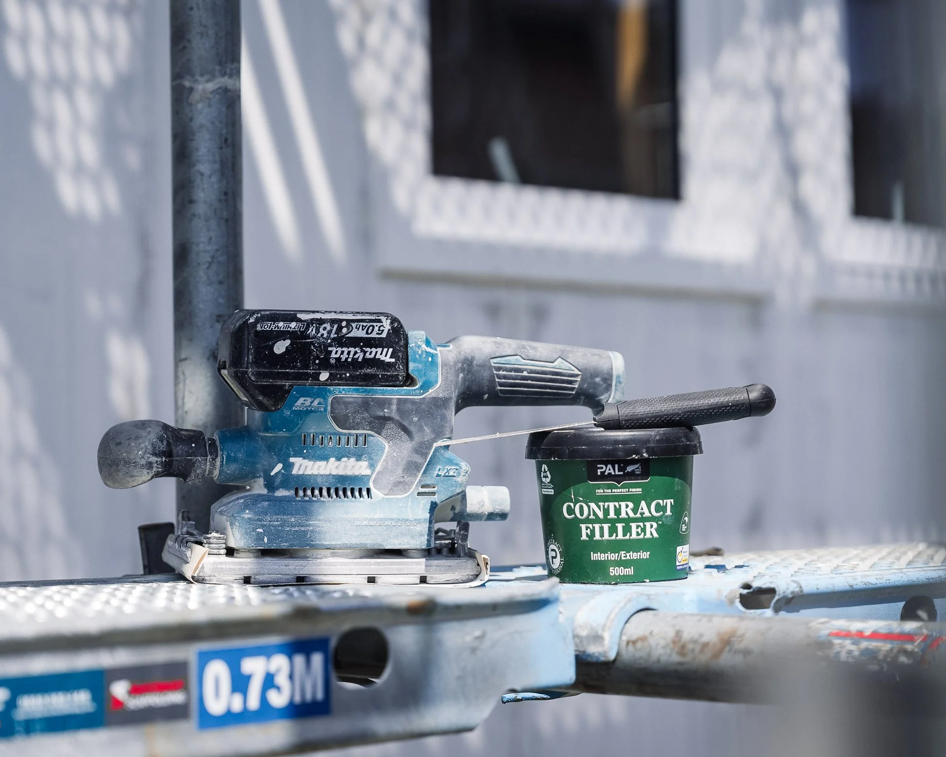 makita orbital sander and pal contract filler pot with putty knife sitting on a scaffold in front of a new build being prepped for painting.