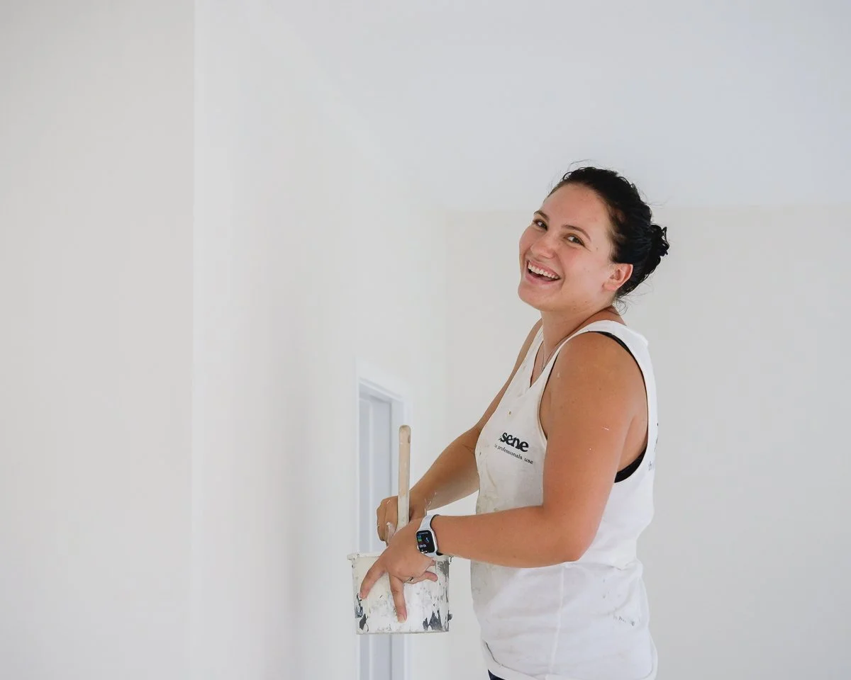 A woman with dark hair in a bun painting a wall white while smiling at the camera.