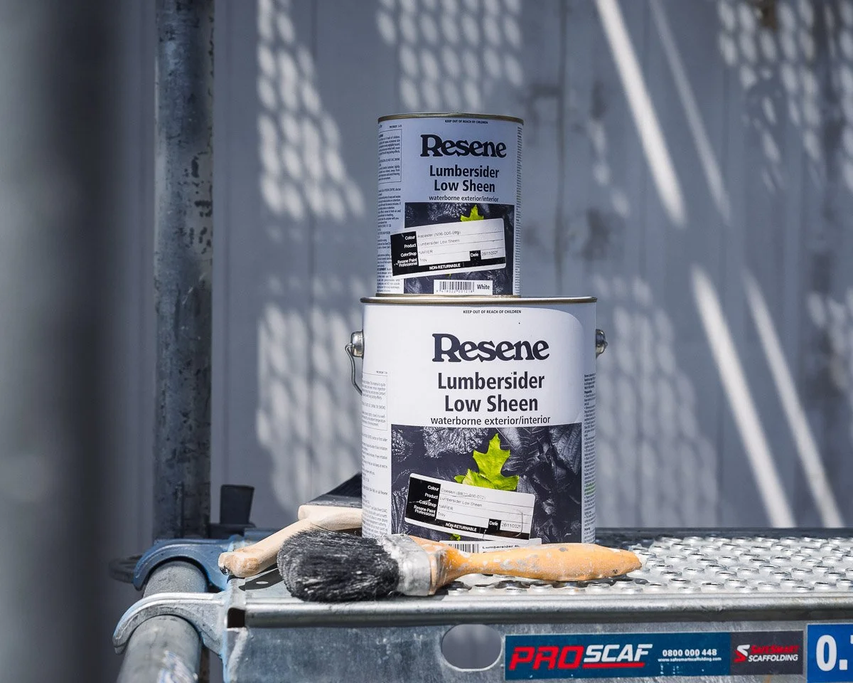 two paint cans sit on scaffolding with paintbrushes, cans are labelled with resene lumbersider exterior paint