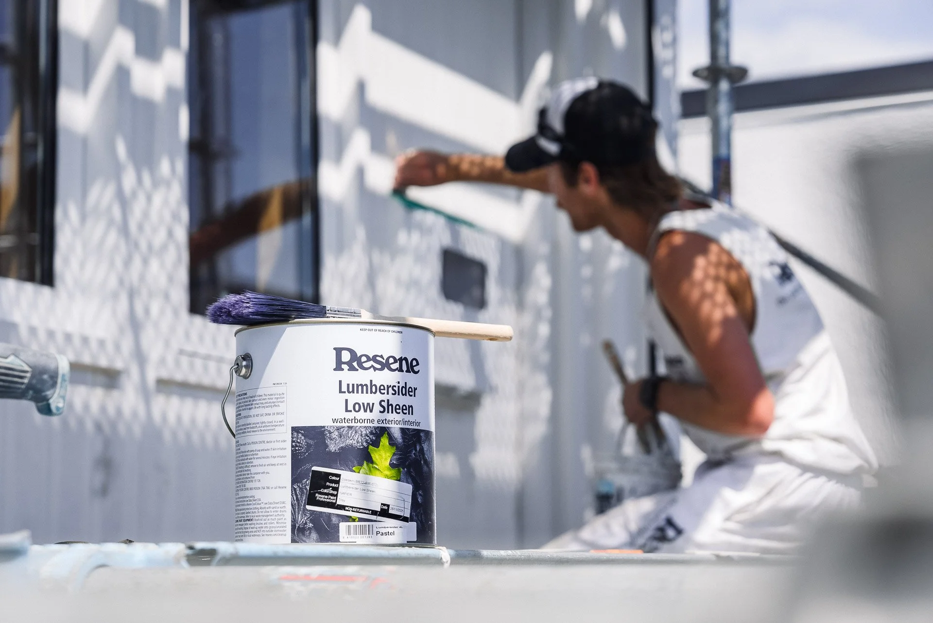 resen lumbersider low sheet exterior paint in the foregroundw ith painter sitting on scaffold painting house cladding with a paintbrush and bucket in backdrop