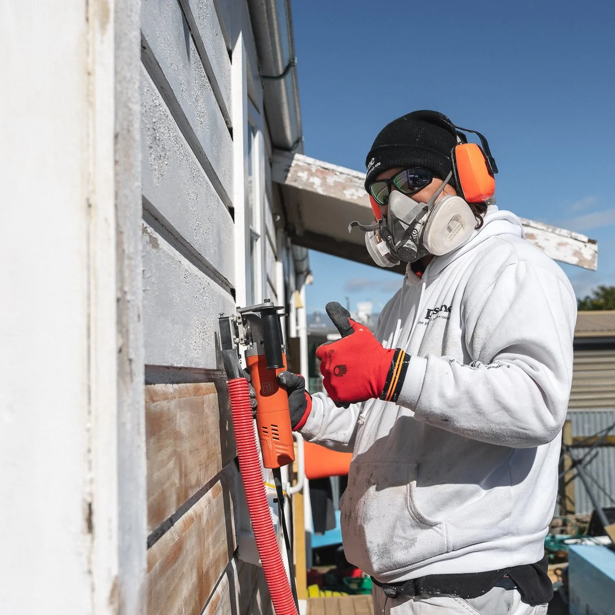 A person wearing protective gear, including a respirator, safety glasses, earmuffs, and gloves, using a paint scraper tool on the exterior weatherboards of an old villa during daytime.