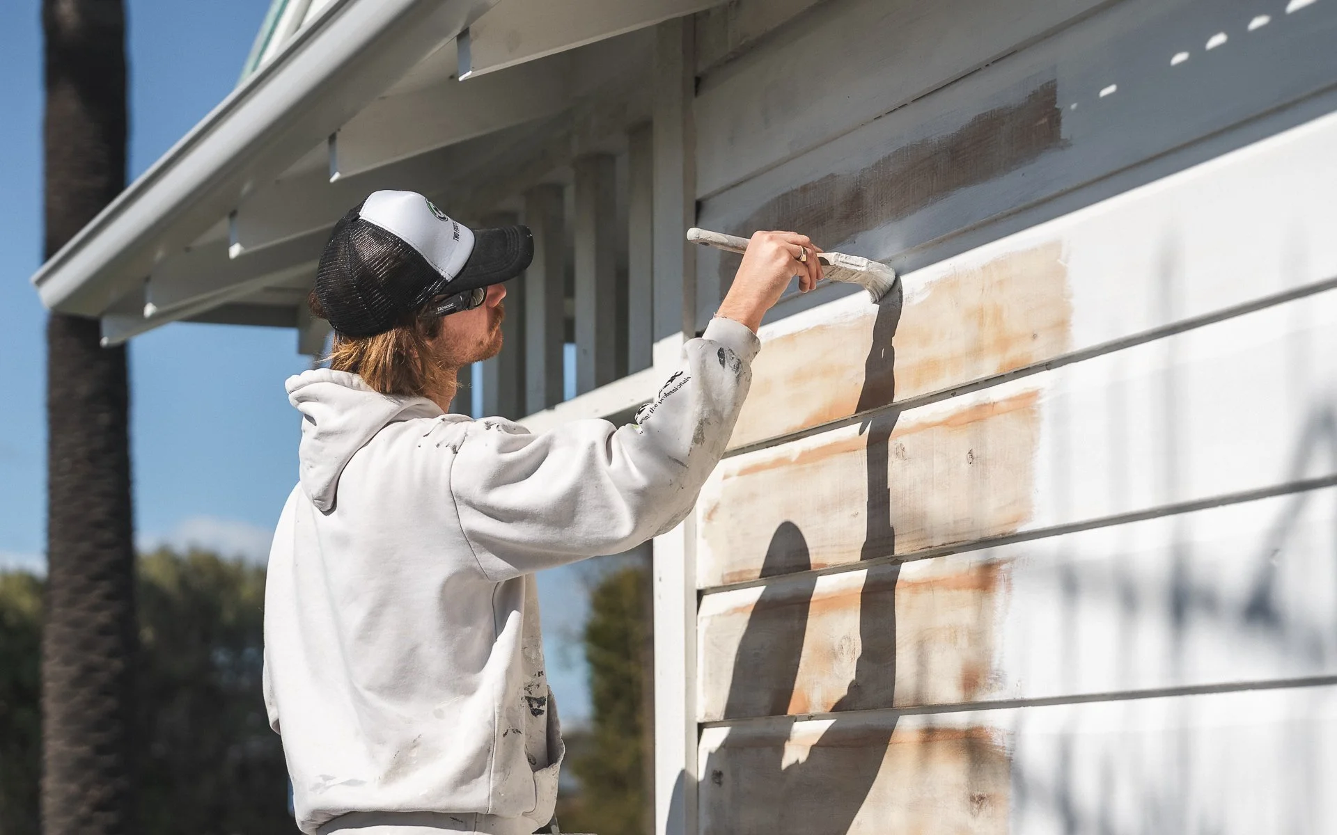 A man wearing white work clothes and a black and white trucker hat is painting the exterior weatherboards of a house with a paintbrush.