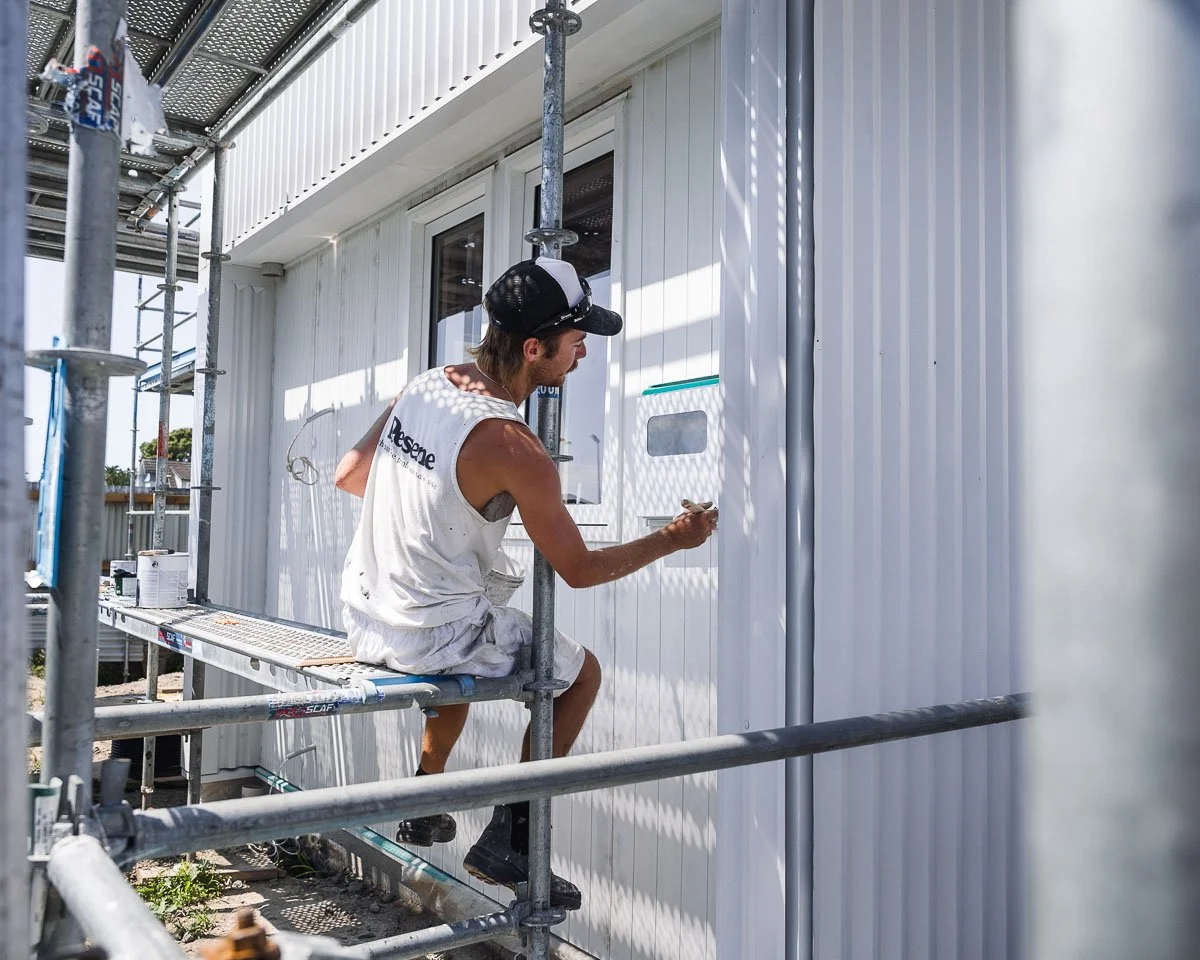 male painter wearing white resene branded painting gear sits on scaffold painting exterior of new build house  in a white colour with a paintbrush
