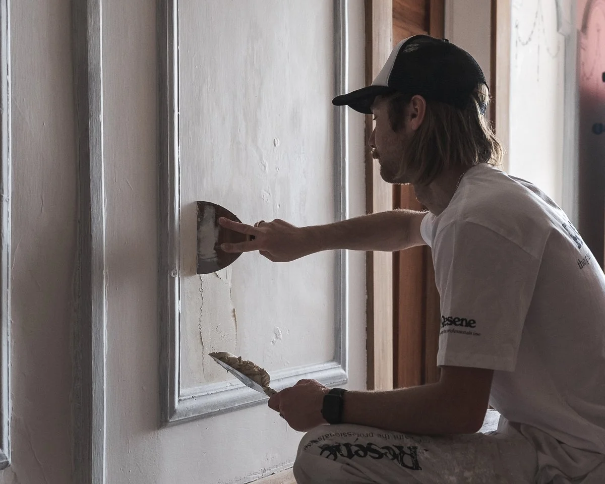 male painter applying plaster to patch a hole on interior wall of character home, he is wearing resene branded painters clothes and a black/white cap