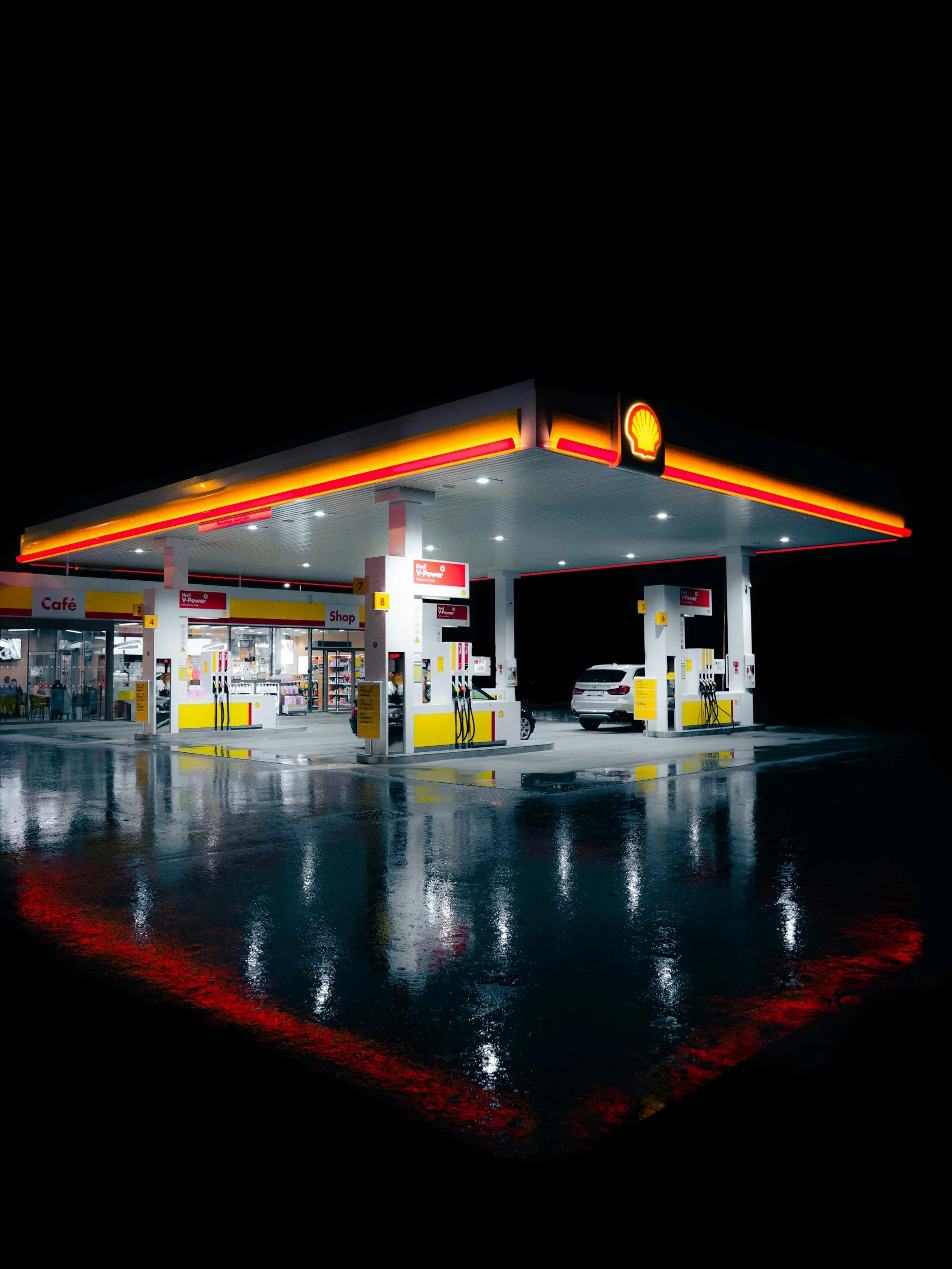 Nighttime photo of a Shell gas station with illuminated canopy and parked cars, reflecting on wet pavement.
