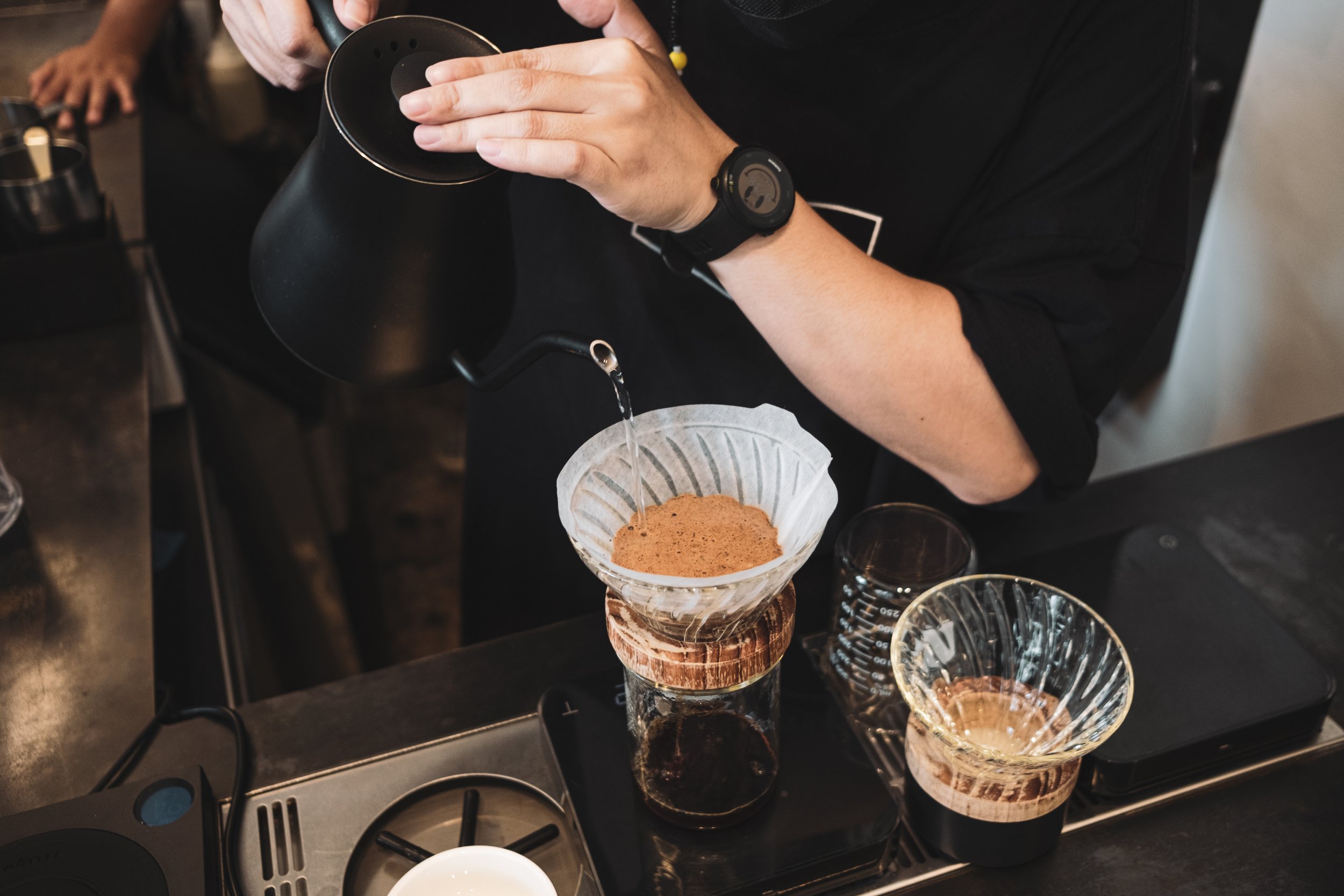 Barista pours hot water over coffee grounds in a paper filter set in a glass dripper, placed on a glass jar. The setup is on a black countertop with coffee brewing equipment around.