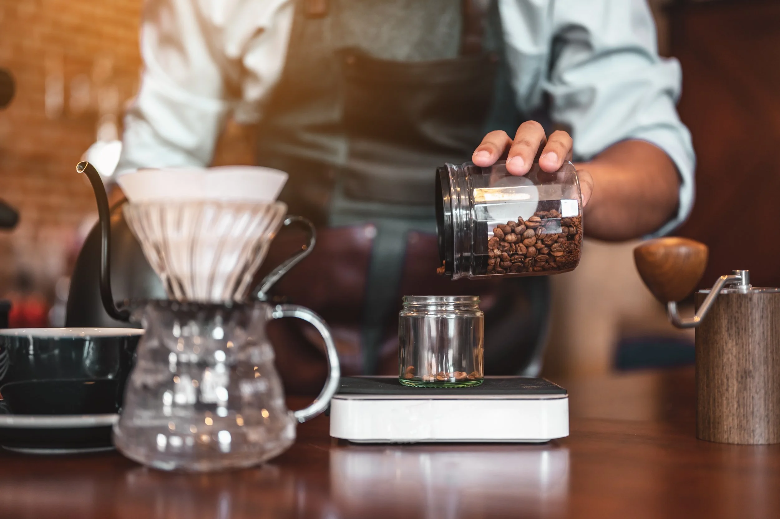 Person pouring coffee beans into a glass container on a scale, with coffee brewing equipment and cups on a wooden table.