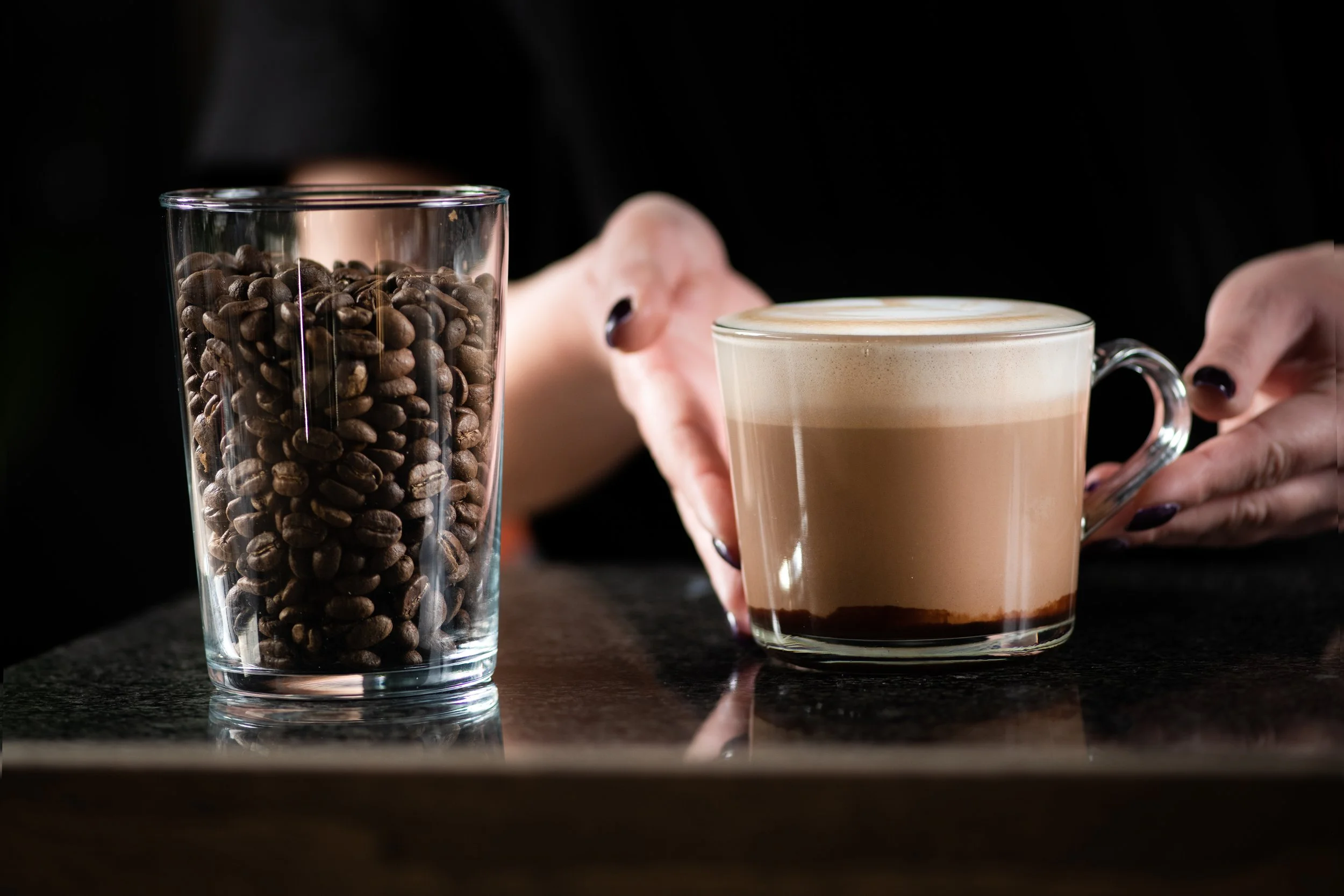 Person reaching for a cup of coffee with milk and foam on top, with a tall glass of coffee beans beside it on a dark surface.