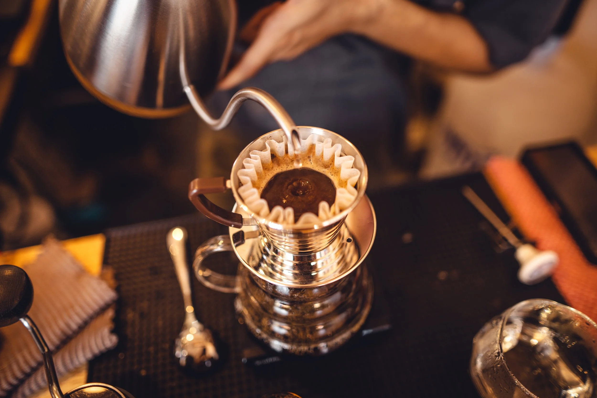 Pour-over coffee brewing process with hot water being poured over coffee grounds in a cone-shaped paper filter, placed on a metallic coffee dripper.