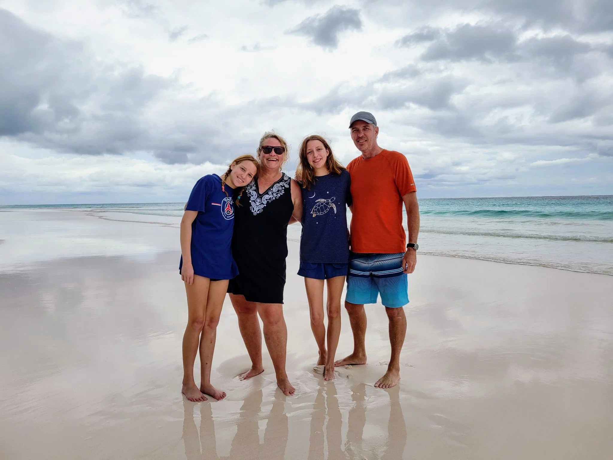 Four people standing on a sandy beach with ocean waves and a cloudy sky in the background, smiling and posing together.