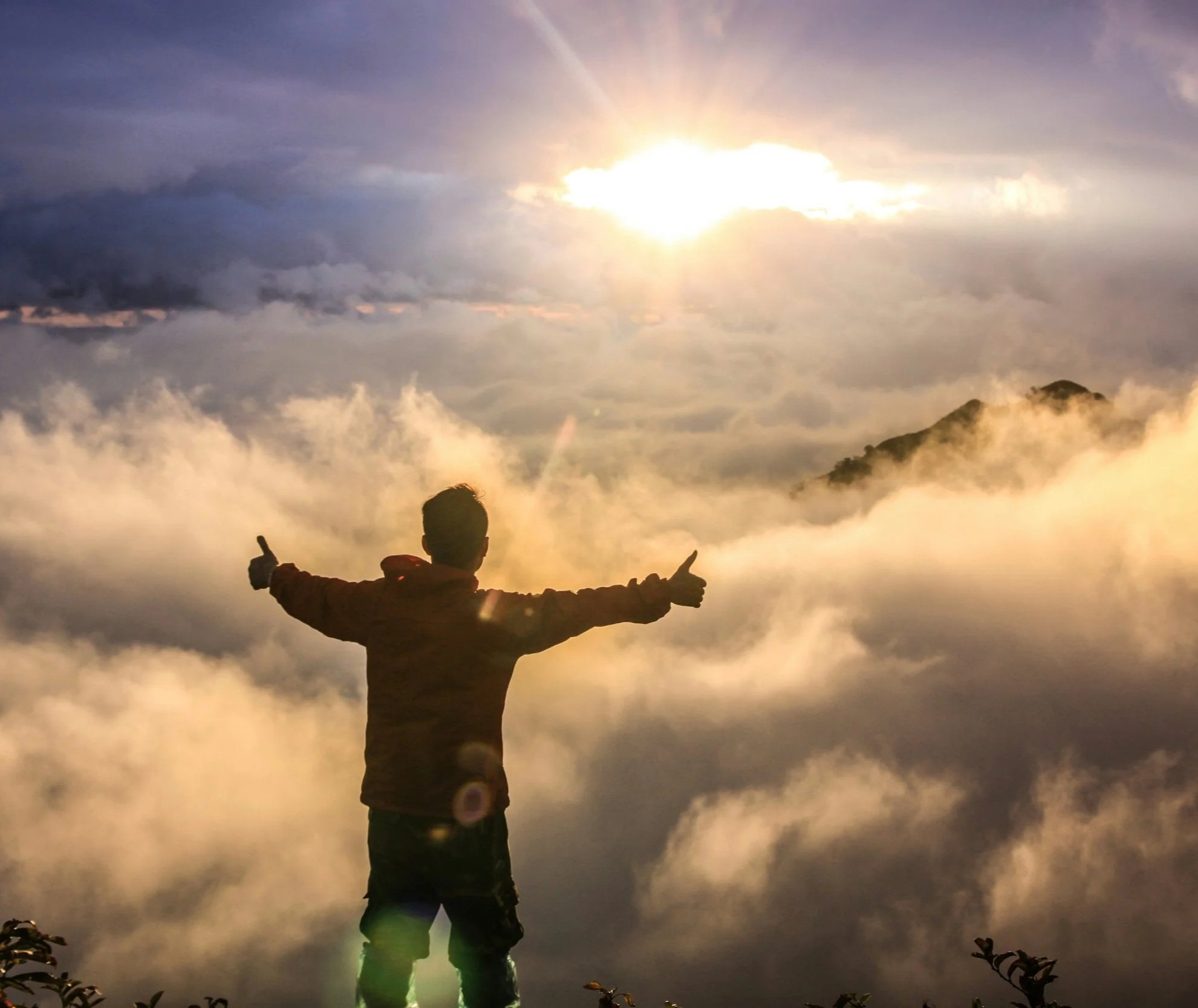 Person standing on a mountain or hilltop with their back facing the camera, arms outstretched, overlooking a landscape of clouds and a mountain range with the sun shining through clouds.