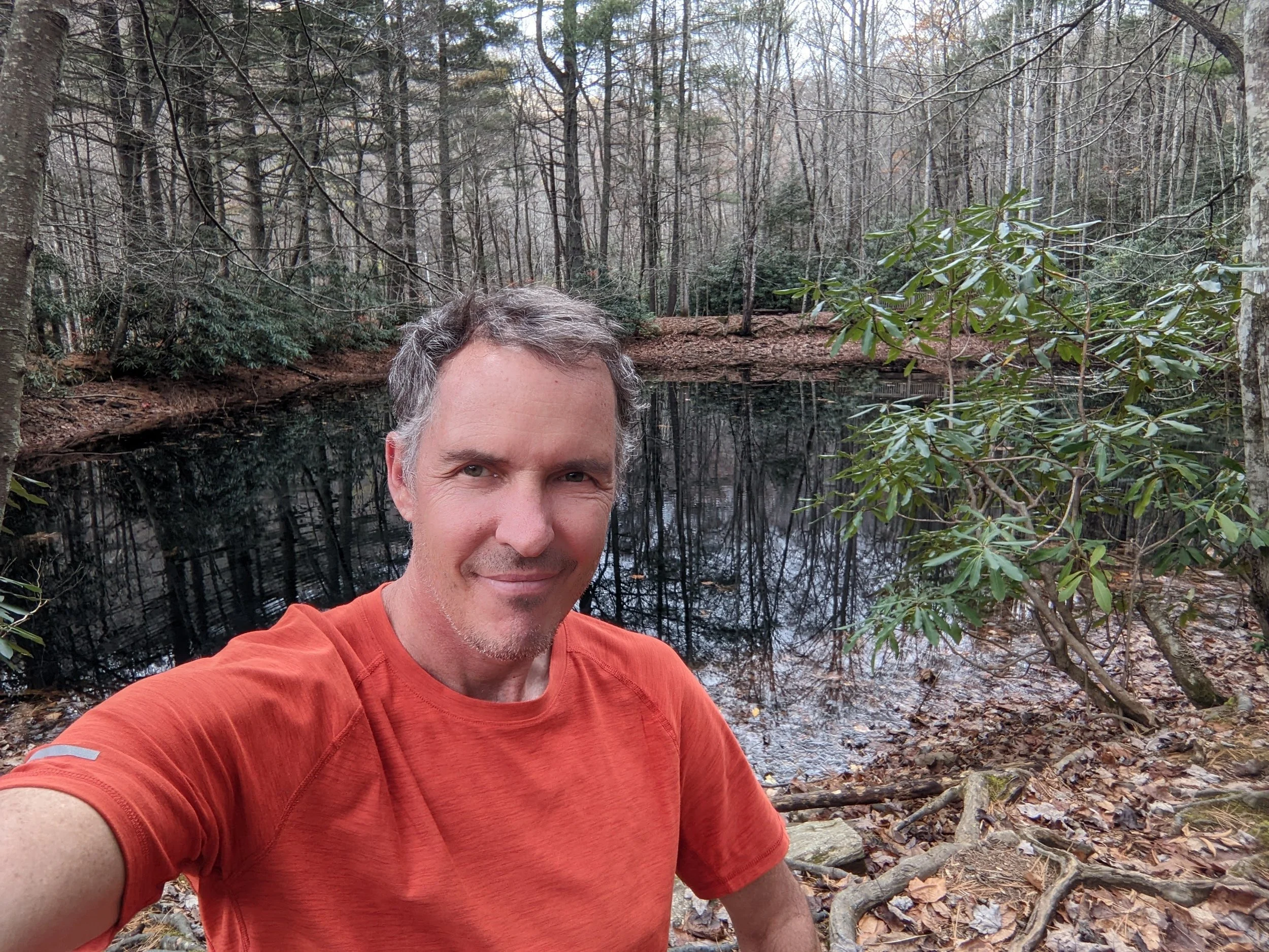 Greg Gillett taking a selfie outdoors in a wooded area near a small pond in Asheville, NC surrounded by trees and fallen leaves.