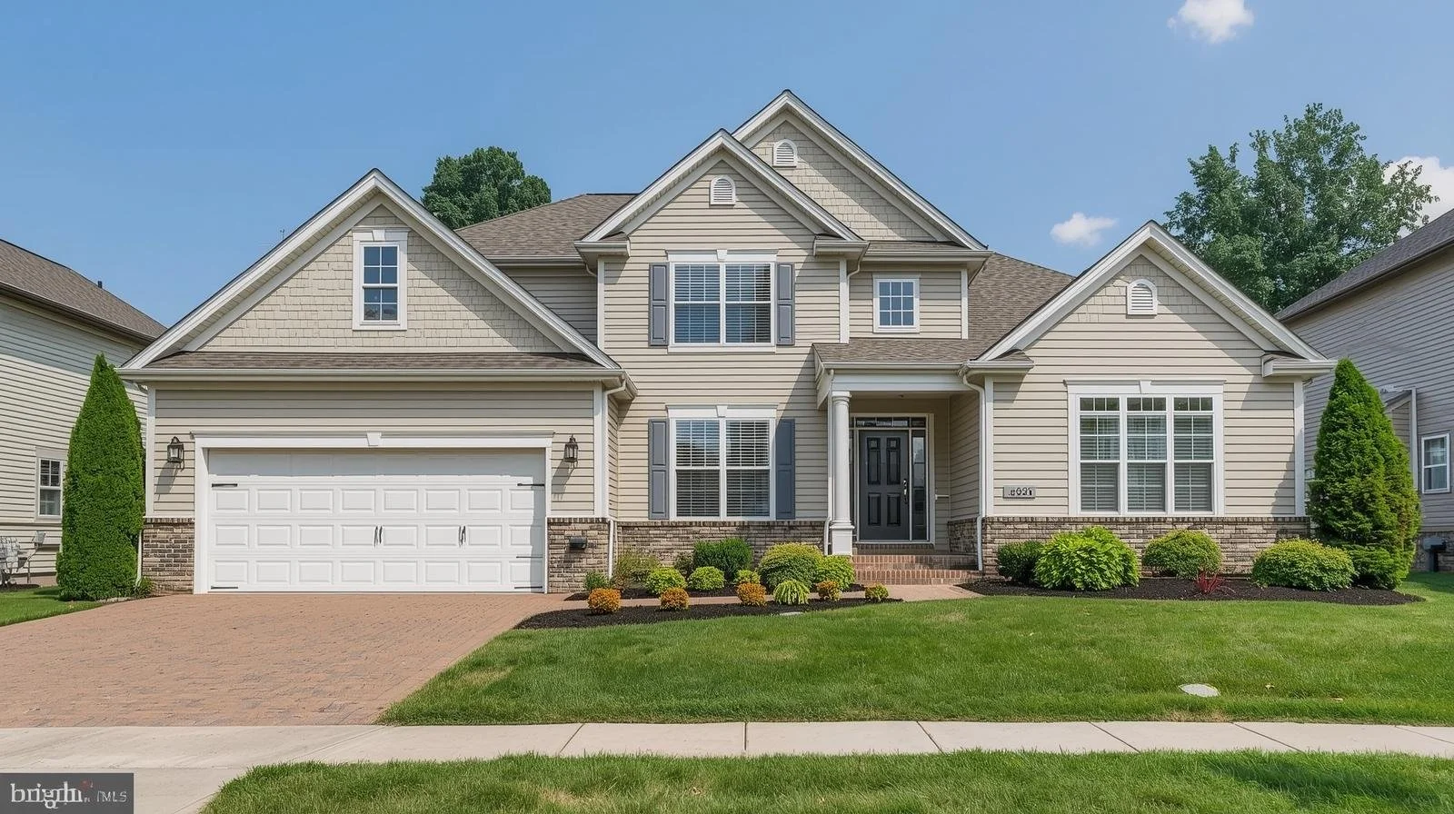Front view of a large suburban house with beige siding, white garage door, manicured lawn, and landscaped bushes and trees.