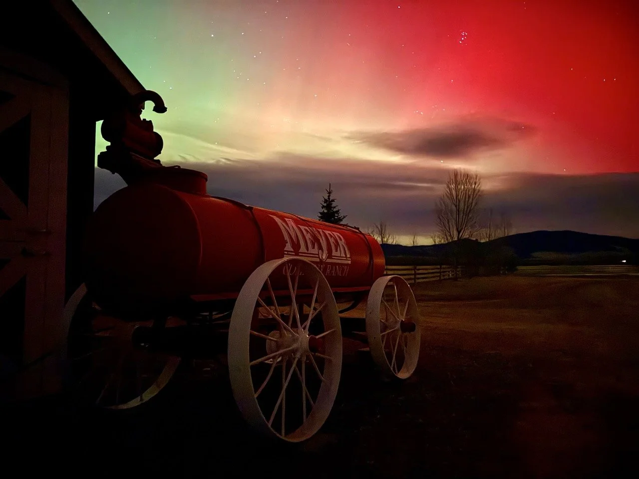 A vintage milk delivery wagon outside a barn with a scenic night sky displaying the Northern Lights and stars in the background.