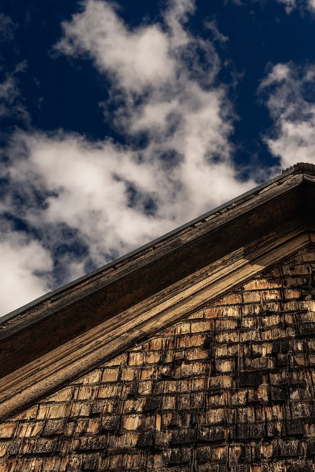 Close-up of a weathered wooden house roof with shingle siding, against a background of dark blue sky with scattered white clouds.