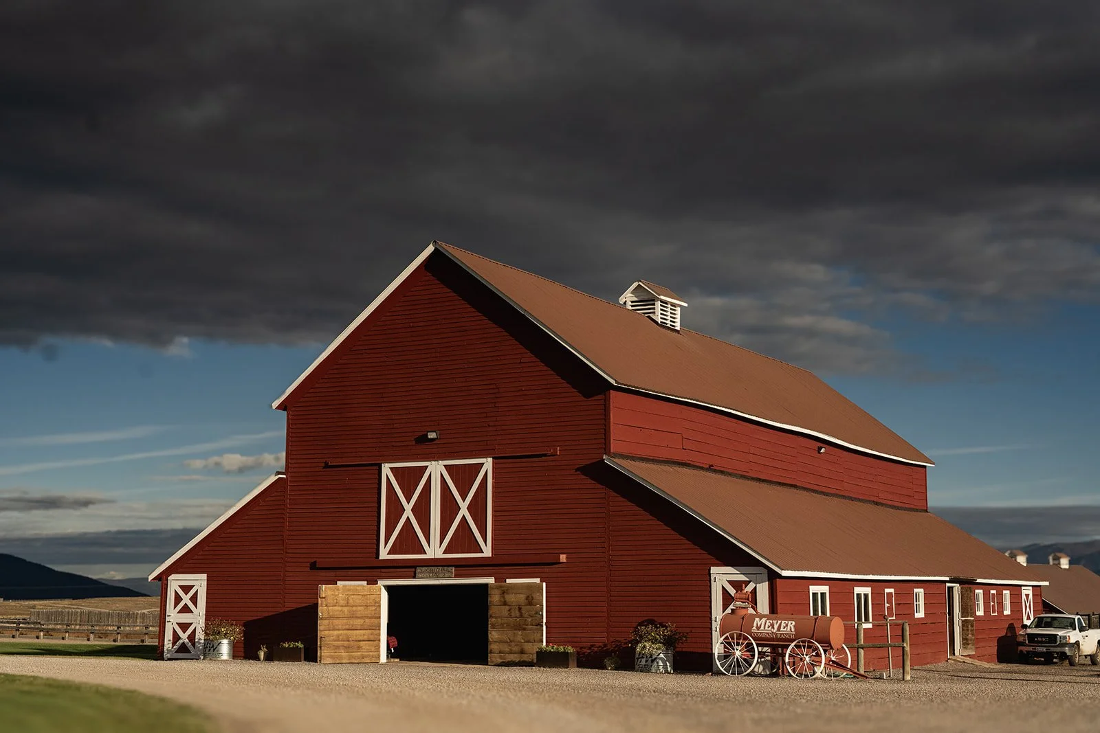 A red barn with white trim, set against a cloudy sky, with a vintage red milk can on wheels in front, and a pickup truck nearby.