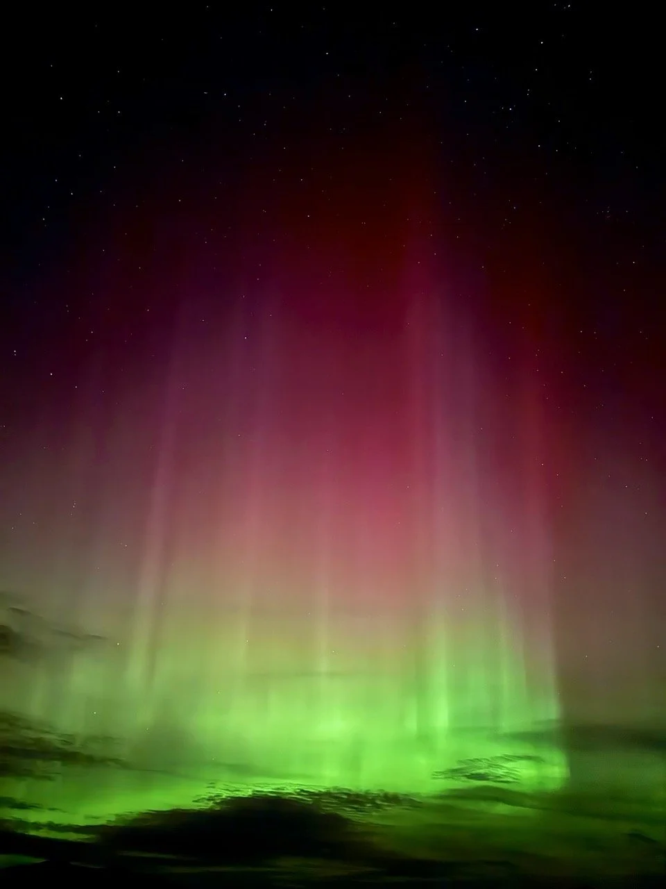 Northern Lights illuminating the night sky with green and red hues, visible stars, and a silhouette of the horizon.