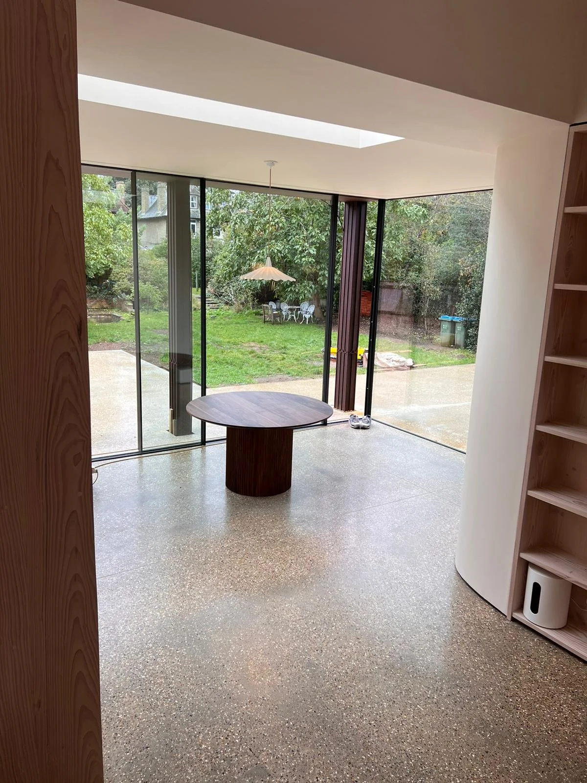 An interior room with large glass sliding doors opening to a backyard with trees and outdoor furniture. A round wooden table is in the center, with shelves on the right side of the room.