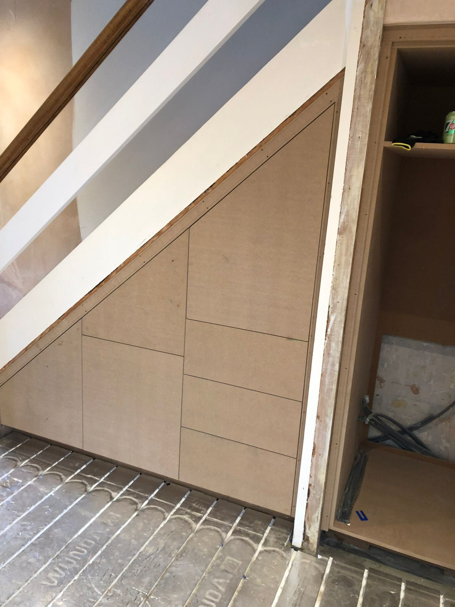 Inside a home under construction, wood framing and drywall are visible, with some drywall installed on a sloped wall next to a staircase. There are electrical wires and tools on the unfinished storage area.