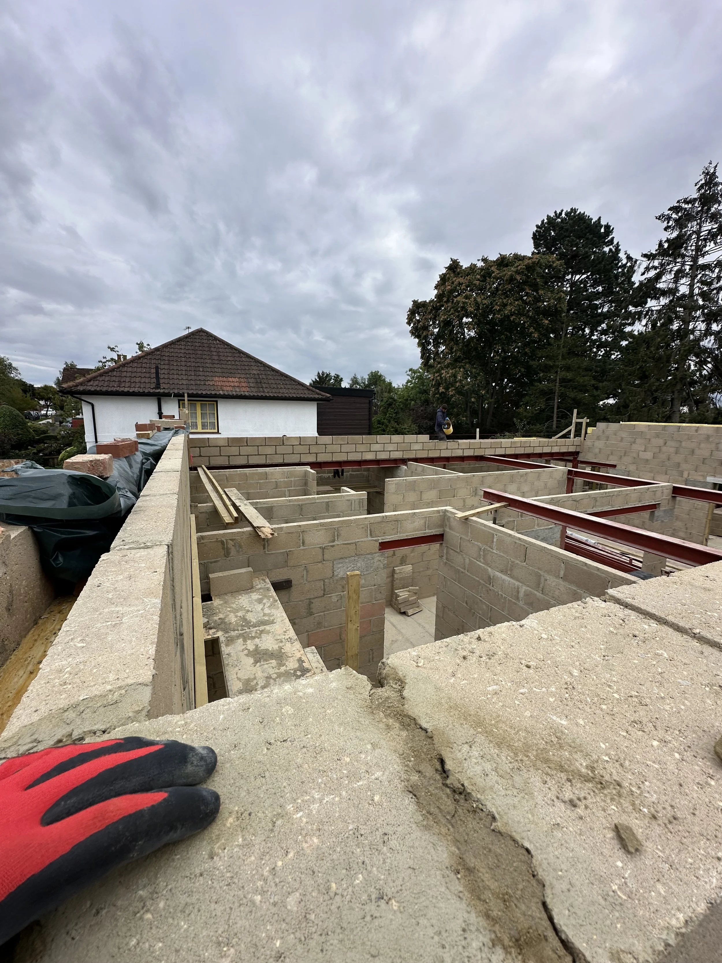 Construction site with partially built brick walls and steel beams. A worker is walking aside, and the sky is cloudy.