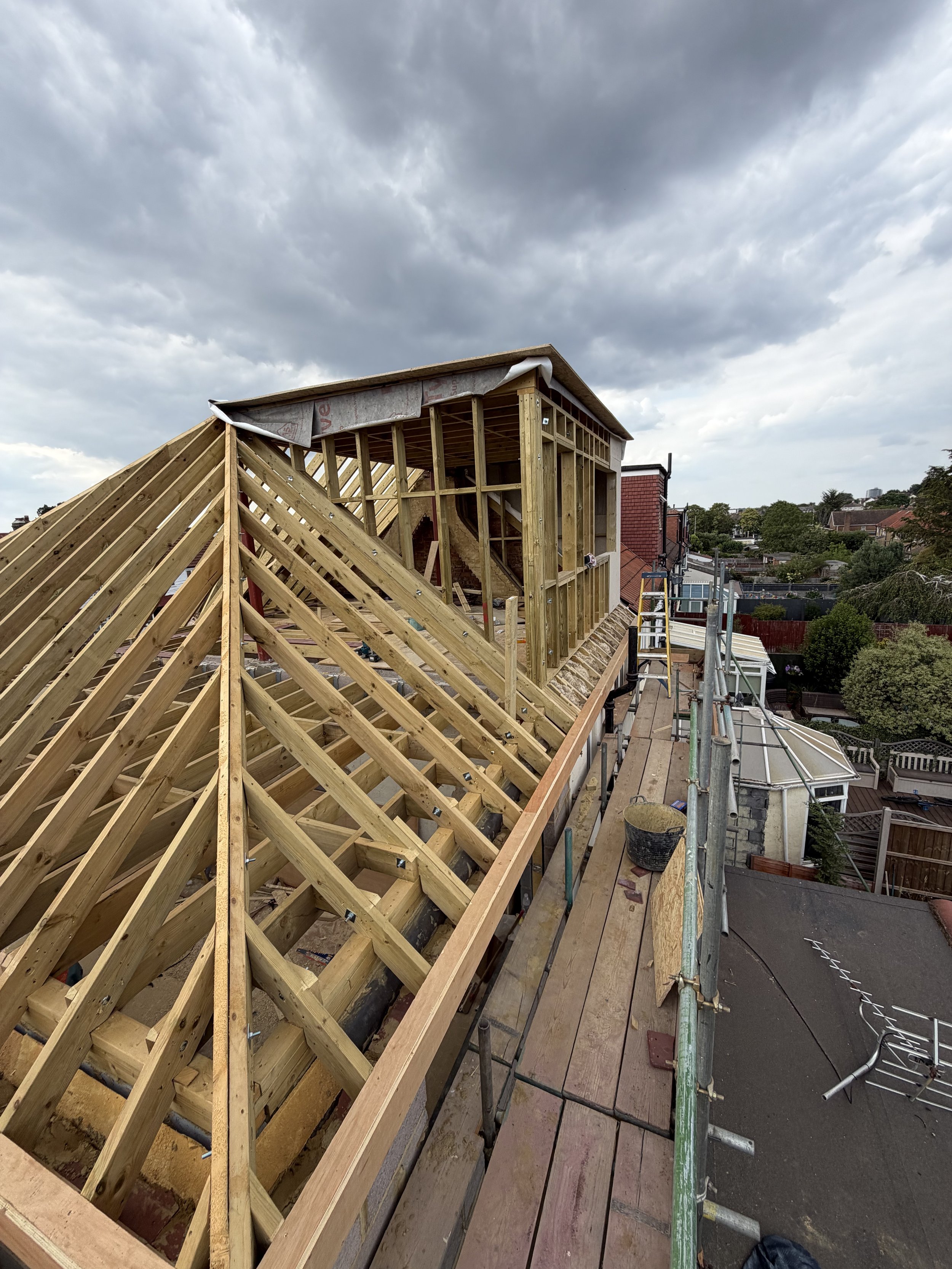 Construction site on the roof of a building showing wooden framing for a new roof structure, with a cloudy sky overhead and urban neighborhood in the background.