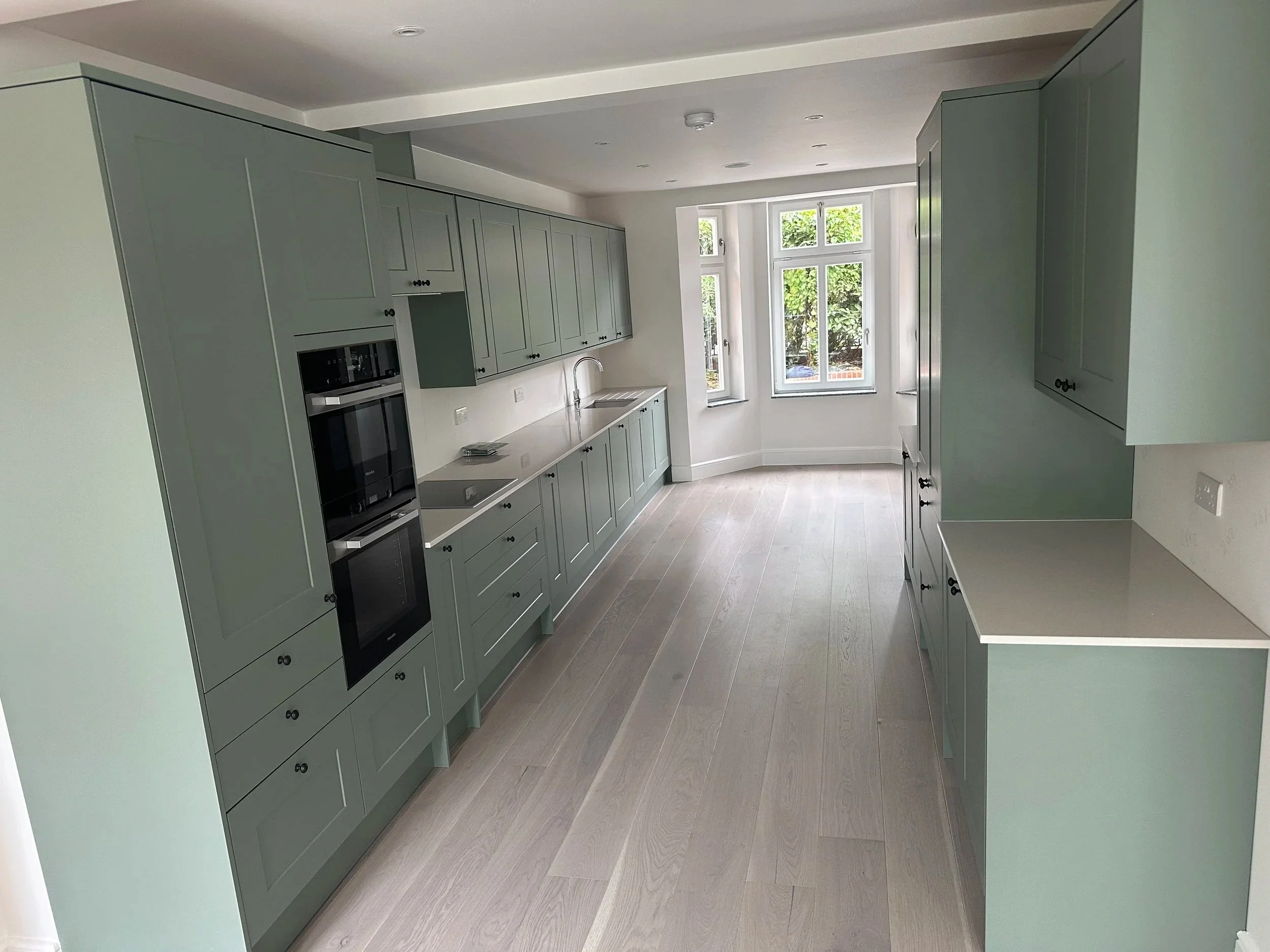 Modern kitchen with mint green cabinets, a double oven, a sink under a window, and light wooden flooring.