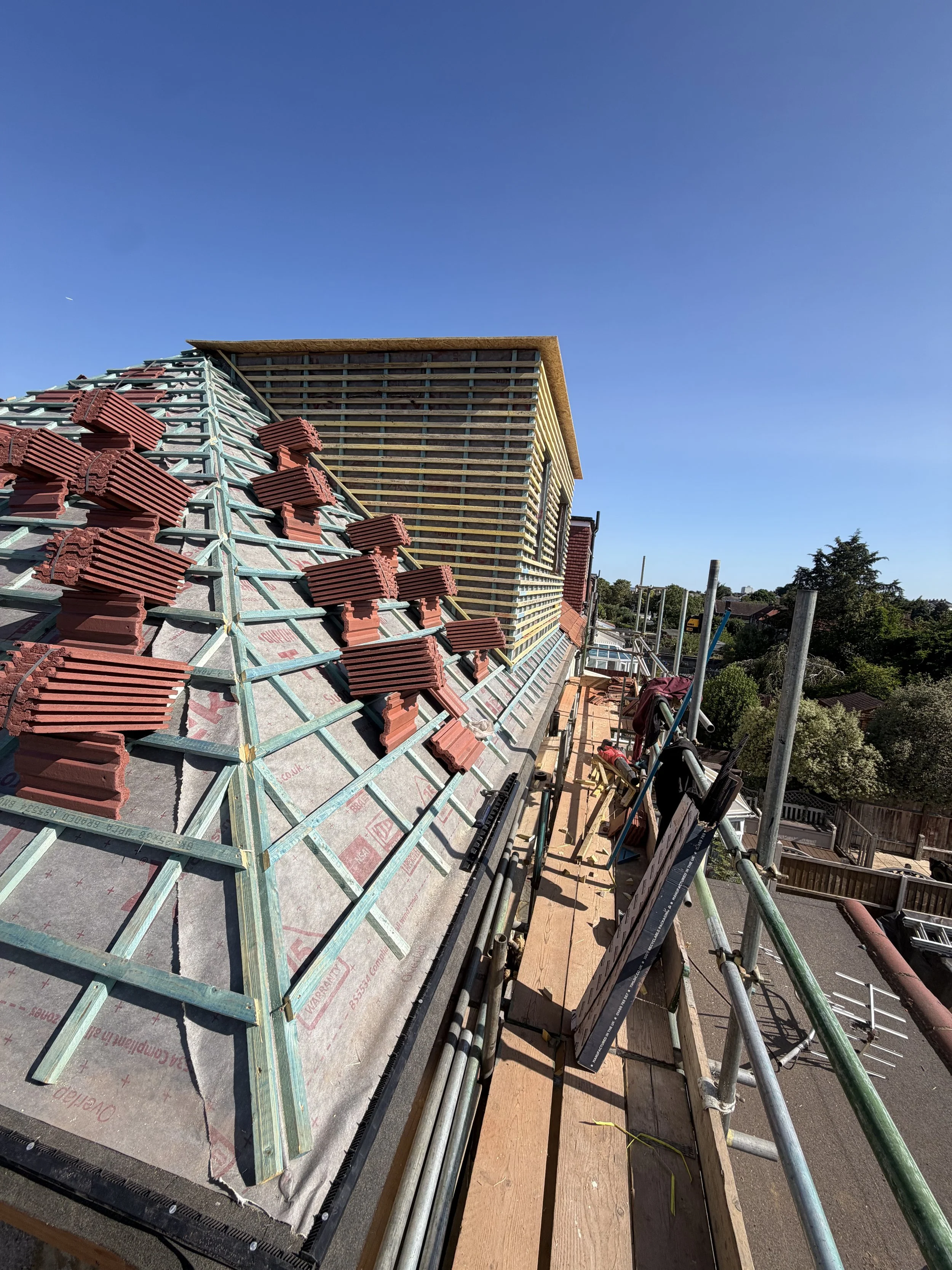 Construction site on a house roof with roofing tiles and wooden framing, with scaffolding and tools on the edge, and trees and houses in the background.